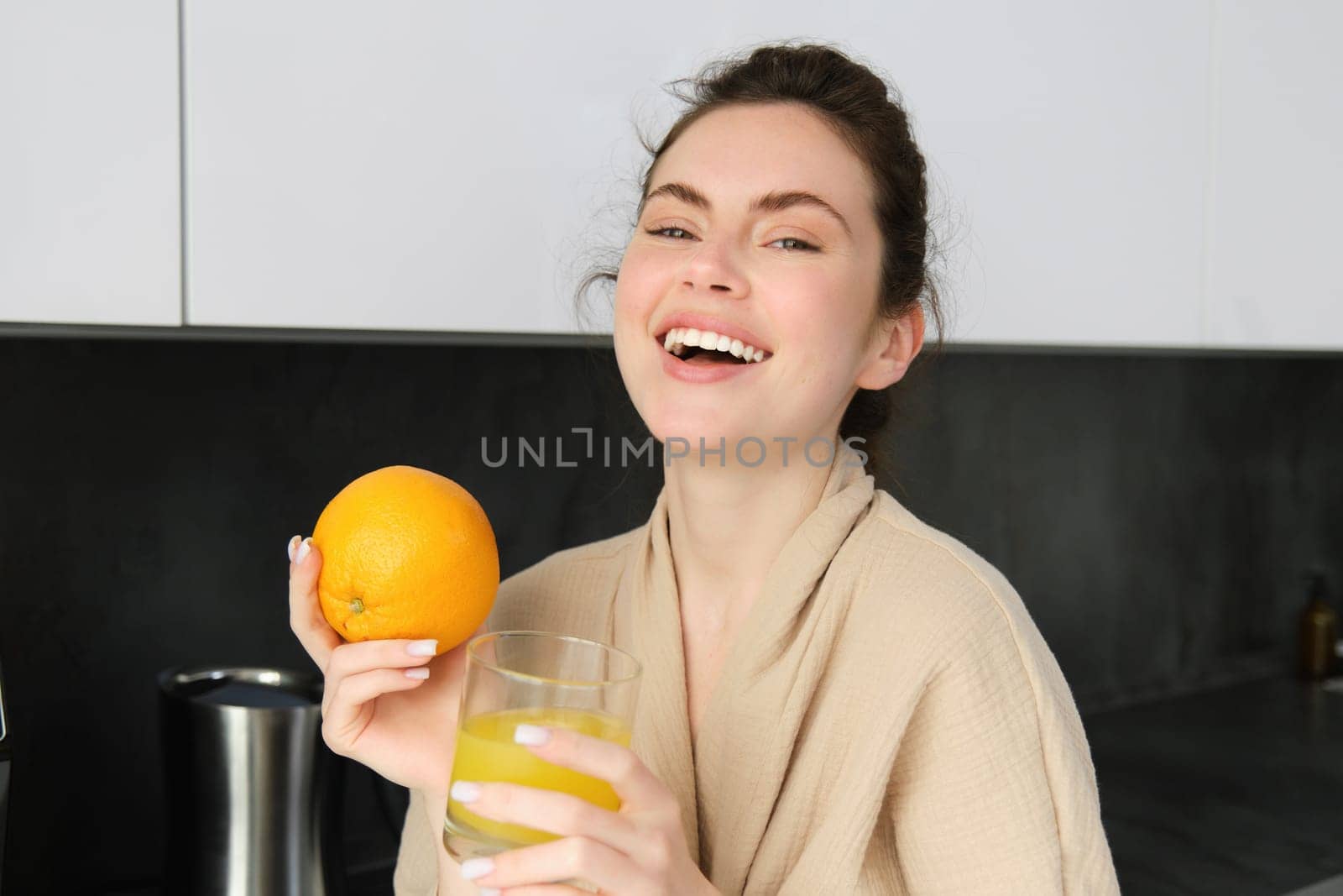 Portrait of beautiful, healthy young woman with orange in hand, drinks freshly squeezed juice from glass, posing in kitchen, enjoys her morning at home.