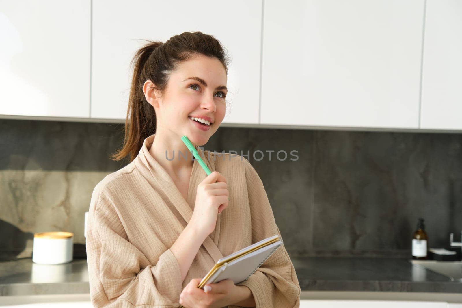 Portrait of beautiful modern woman, writing down grocery list, meal ideas in notebook, standing in the kitchen, wearing cosy bathrobe.