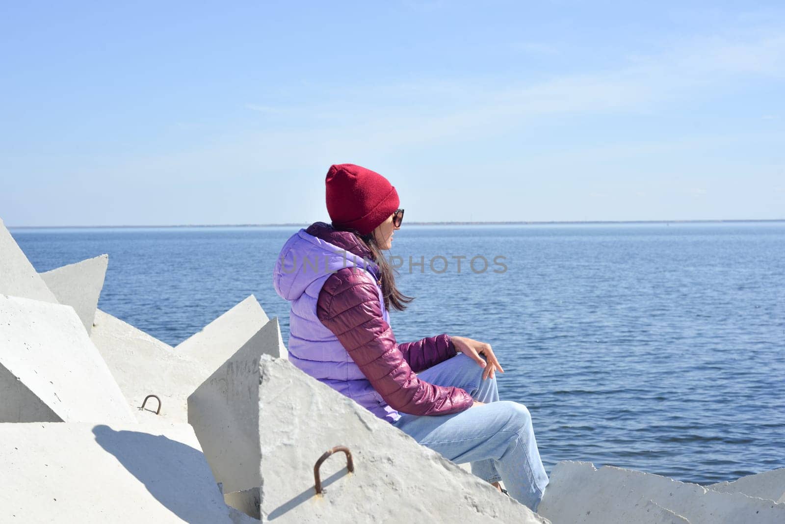 A young woman in a hat on a sunny spring day sits on the breakwaters and admires the sea. Girl in warm clothes on the breakwater