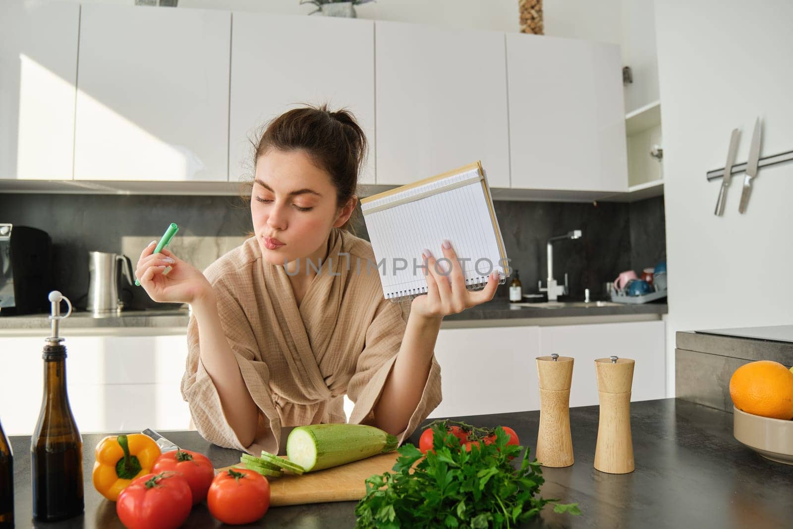 Portrait of modern young woman cooking, making grocery list, reading recipe and making meal, salad in the kitchen, looking at vegetables on chopping board by Benzoix