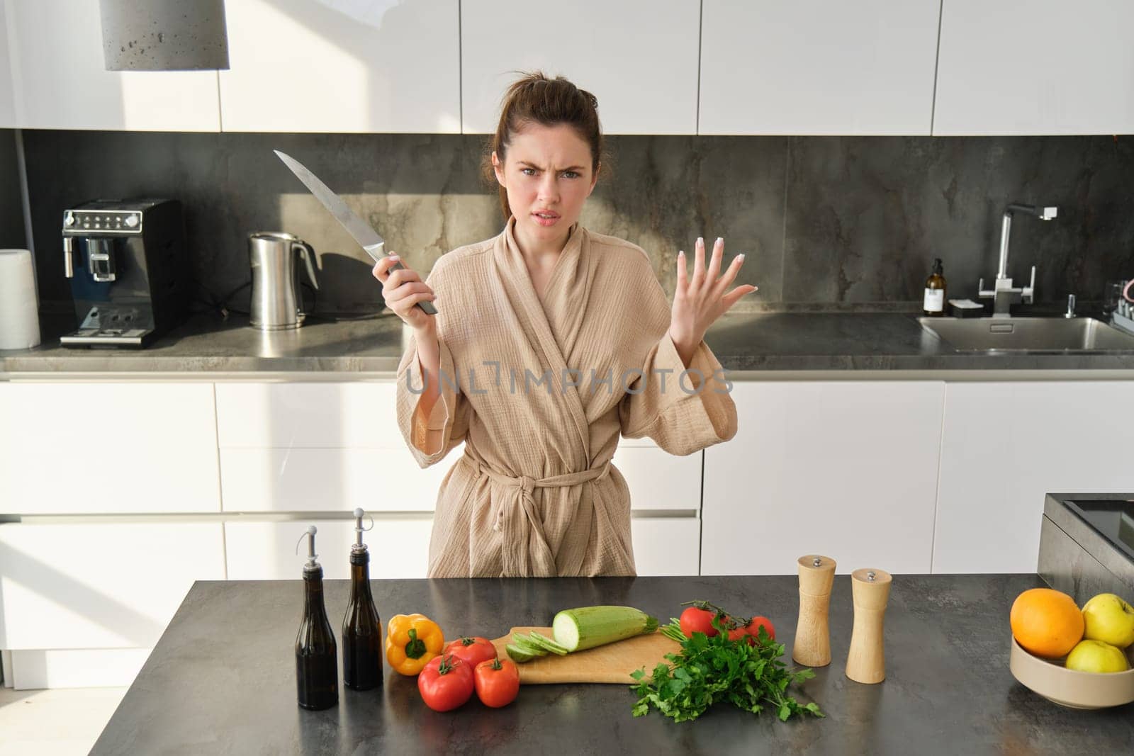 Portrait of annoyed woman with knife, angry while cooking in the kitchen, frustrated while doing house chores and preparing food for family, standing in bathrobe by Benzoix