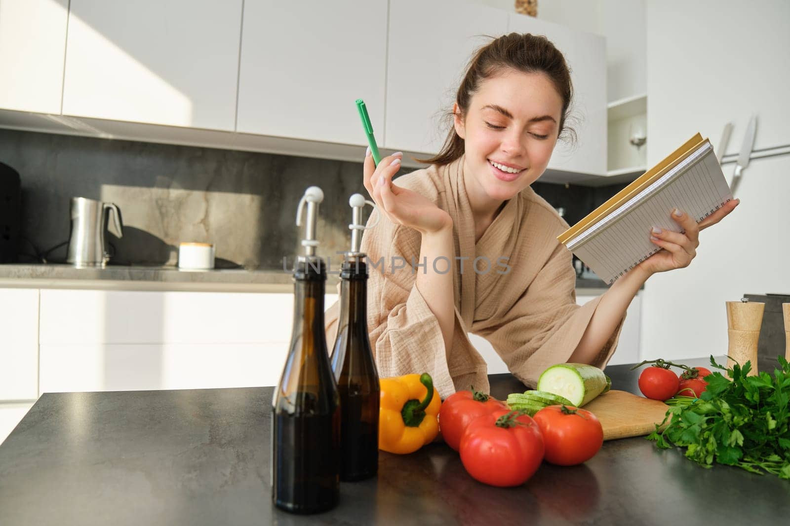 Portrait of woman checking recipe notes in notebook, standing in kitchen with vegetables, cooking food, preparing delicious salad from tomatoes, parsley and olive oil.
