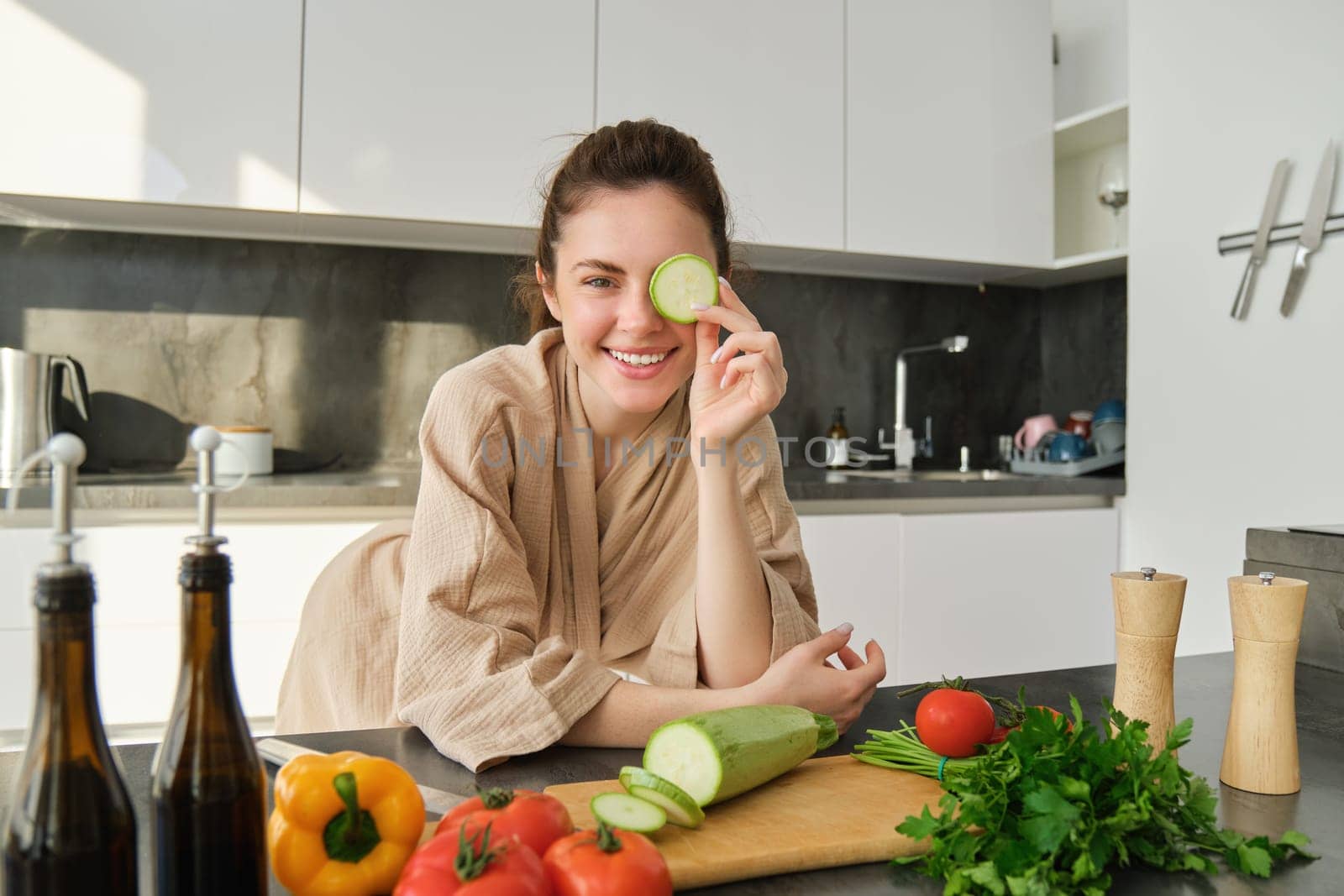 Portrait of smiling brunette woman, girl cooks vegetables, posing with peace of zucchini, leans near chopping board and looking happy while preparing meal, cooking healthy food.