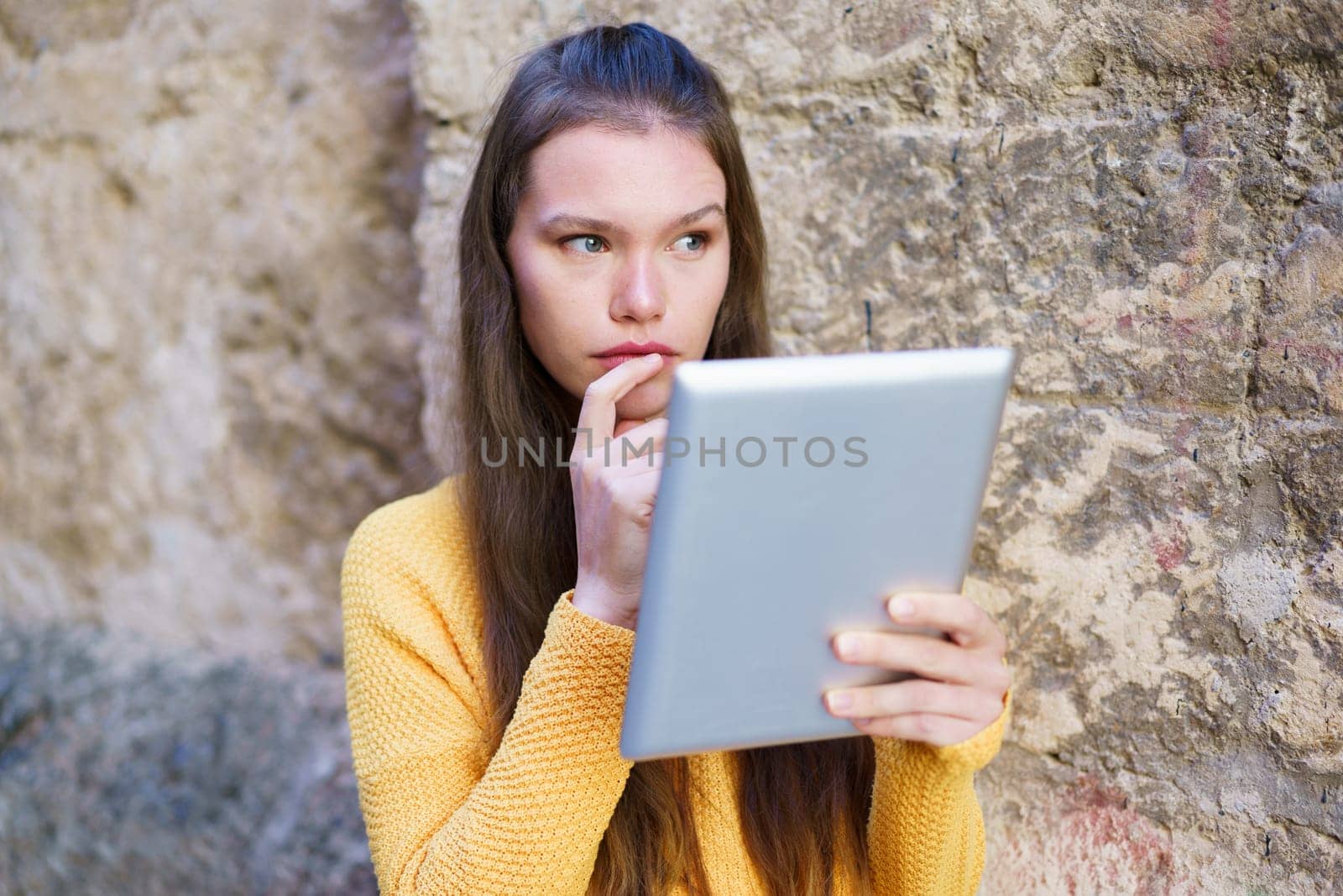 A young woman in a bright yellow sweater engages with her tablet while leaning against a rustic stone wall, blending contemporary tech with deep contemplation and personal reflection