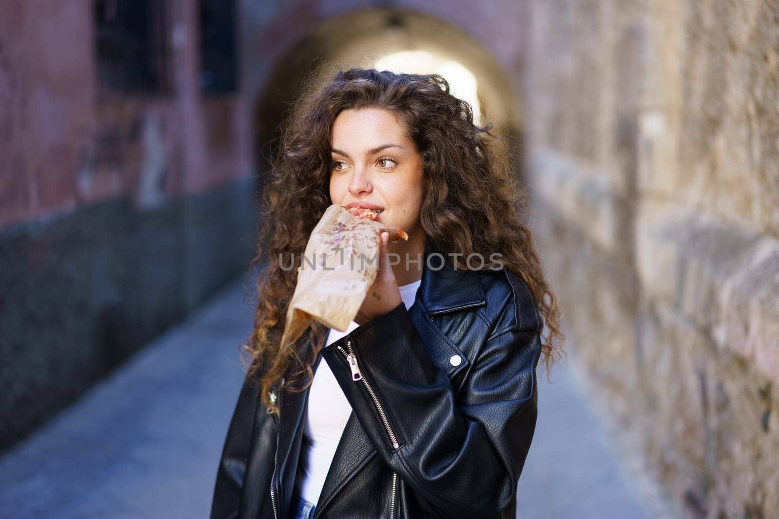 In a vibrant urban alley, a spirited young woman with lively curly hair, sporting a stylish leather jacket, indulges in a delicious snack, fully embracing a relaxed lifestyle admired by many others
