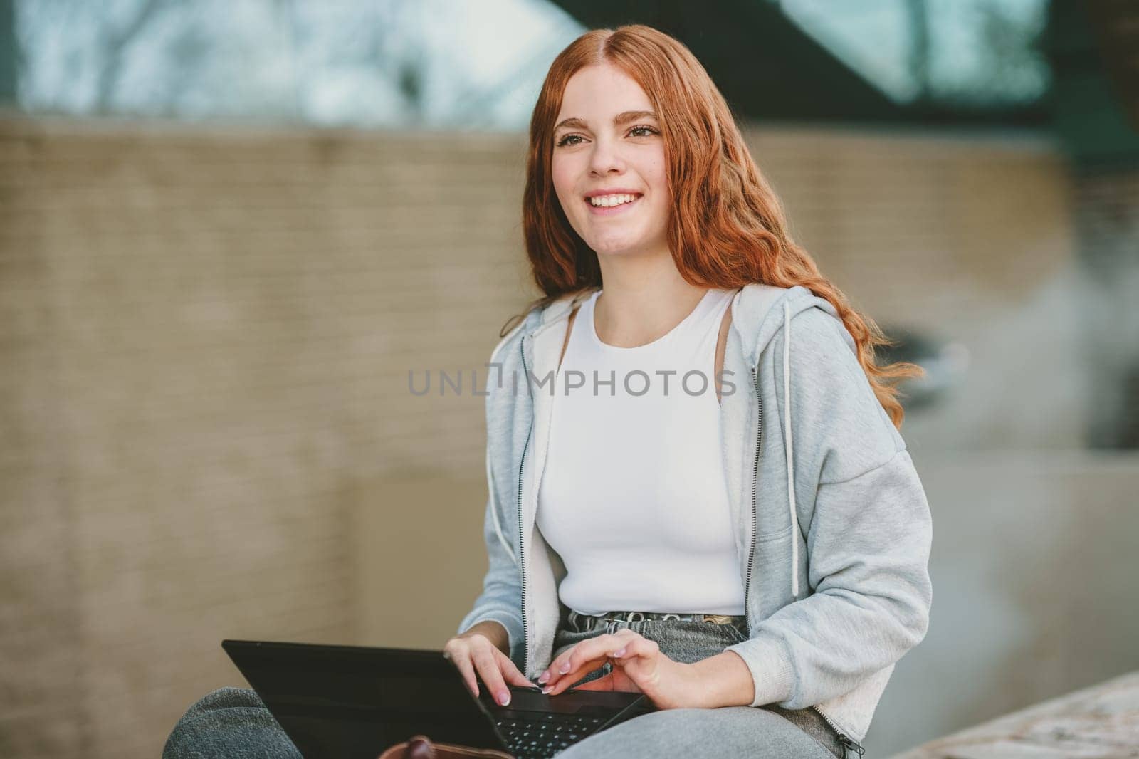 A young woman with long red hair sits outdoors with a smile, working on her laptop. She radiates positivity and productivity in a vibrant setting, reflecting her carefree yet focused lifestyle