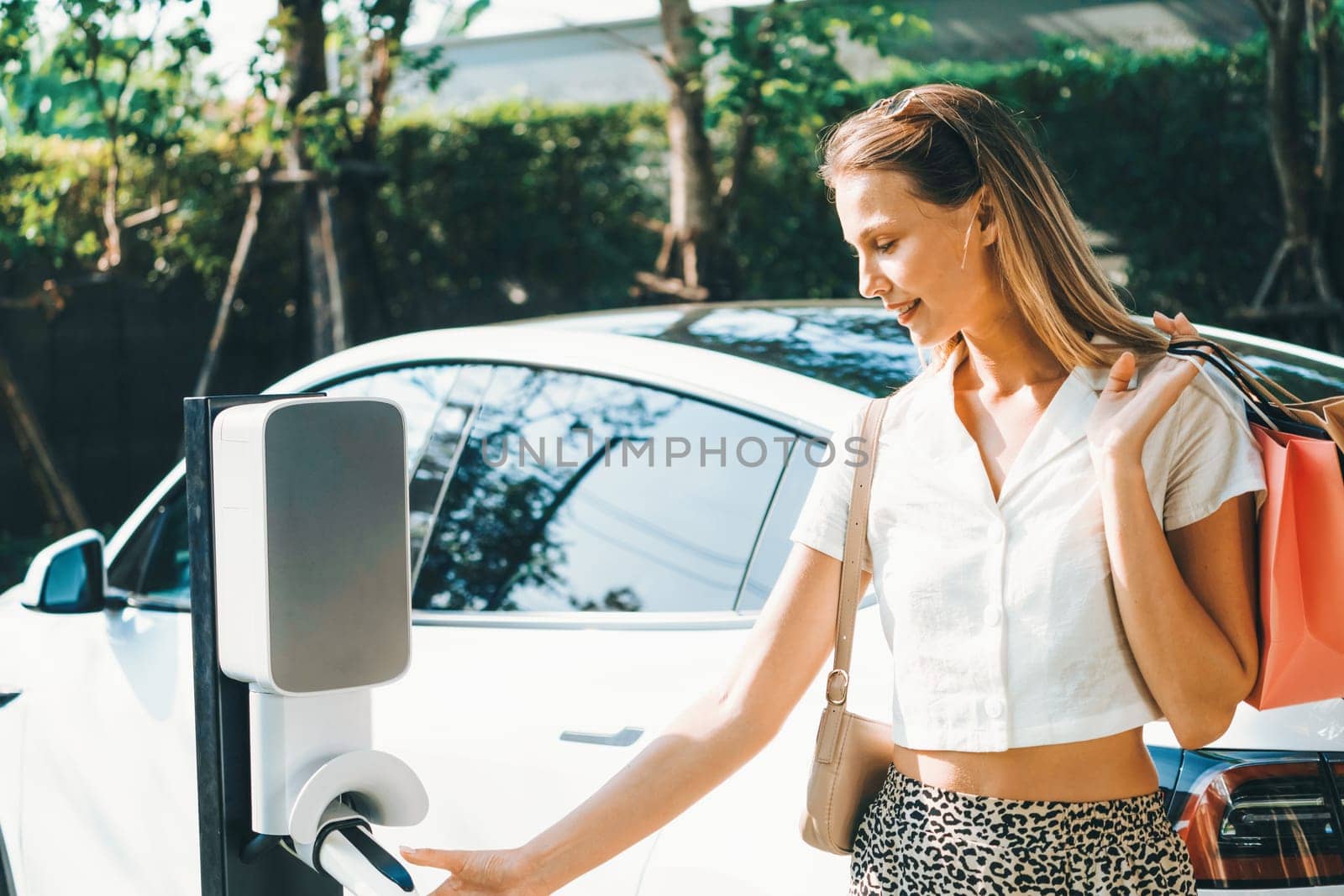 Young woman travel with EV electric car charging in green sustainable city outdoor garden in summer. Urban sustainability lifestyle by green clean rechargeable energy of electric BEV vehicle innards