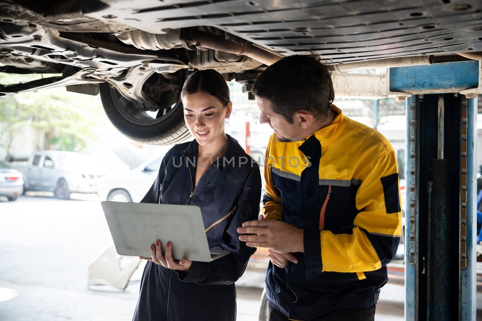 Two vehicle mechanic working together underneath lifted car, conduct car inspection with laptop. Automotive service technician in uniform carefully make diagnostic troubleshooting. Oxus