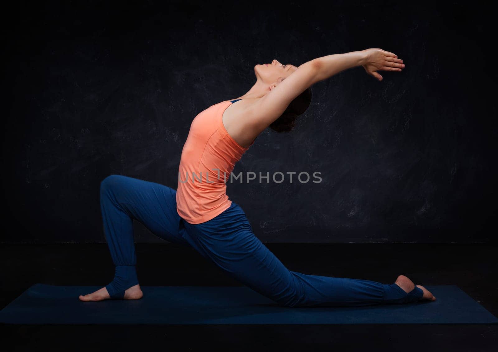 Beautiful sporty fit yogini woman practices yoga asana Anjaneyasana - low crescent lunge pose in surya namaskar on dark background