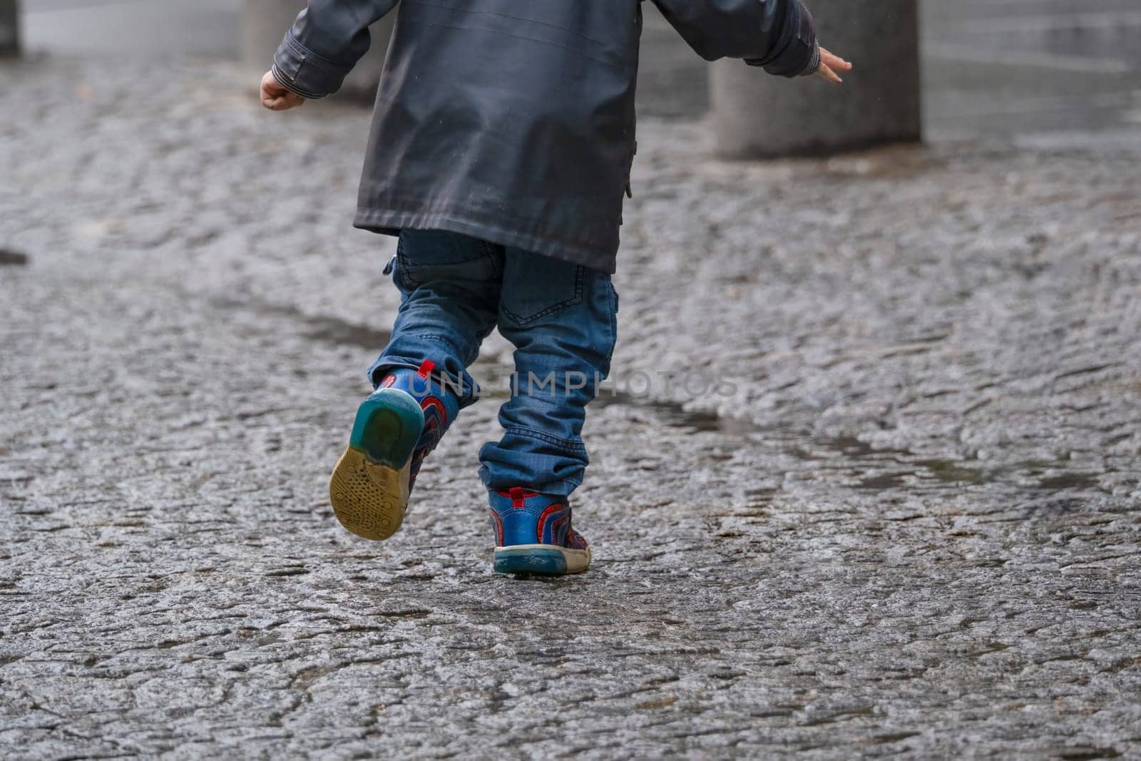Little boy running in rain on urban historic cobblestone street. Arms in position of wings to fly. Copy space. High quality photo