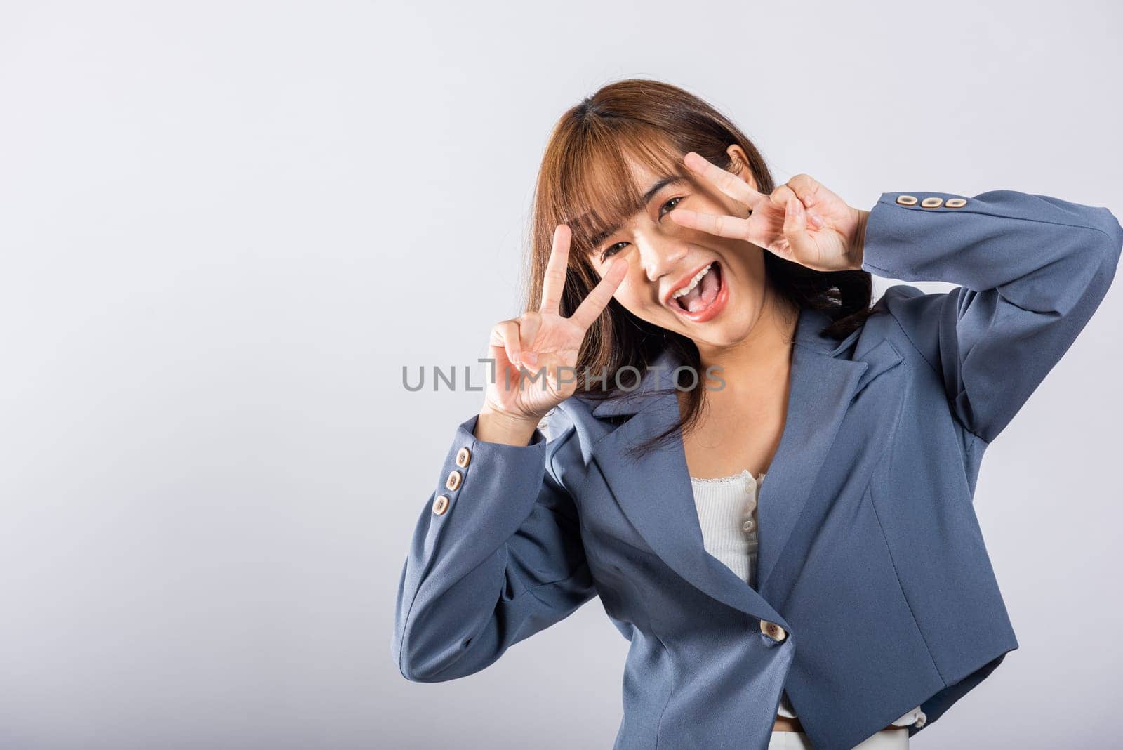 A young Asian woman, photographed in a studio, creatively uses her fingers to display victory and peace signs near her eye. The white background provides a clean canvas for your message.