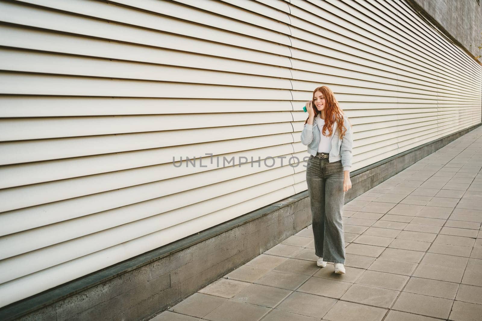 A stylish woman walks a modern sidewalk, focused on her phone, surrounded by sleek architecture. She represents leisure and individuality in trendy jeans and sneakers, enjoying the vibrant city