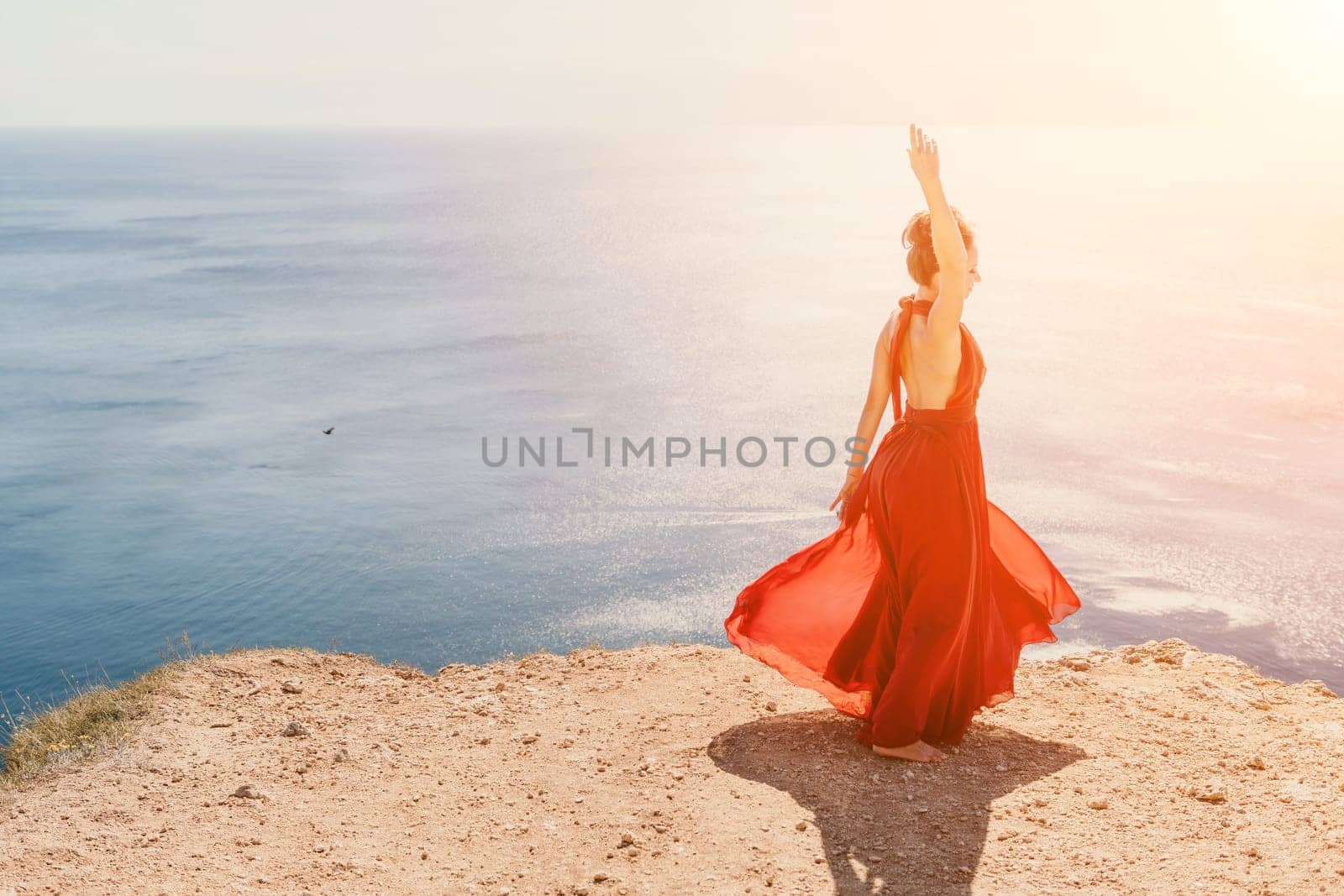 Side view a Young beautiful sensual woman in a red long dress posing on a rock high above the sea during sunrise. Girl on the nature on blue sky background. Fashion photo.