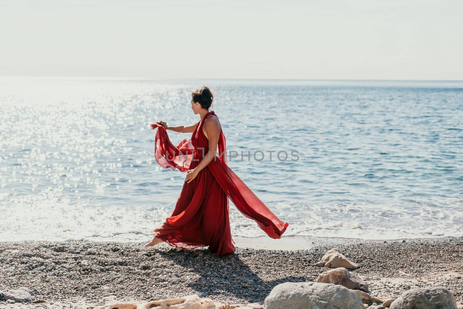 Woman in red dress on sea. Side view a Young beautiful sensual woman in a red long dress posing on the beach near sea on sunset. Girl on the nature on blue sky background. Fashion photo. by panophotograph