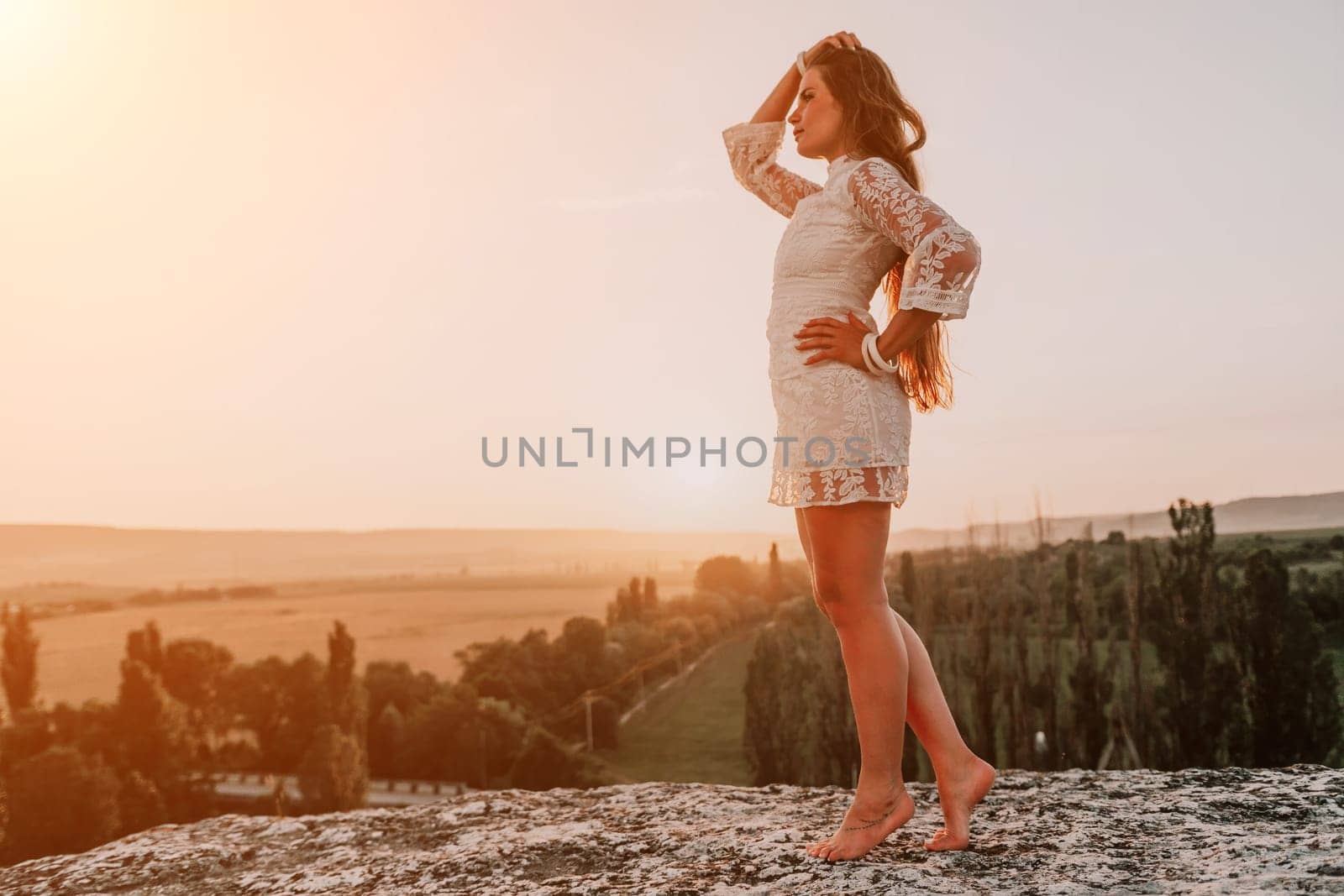 Romantic beautiful bride in white dress posing with sea and mountains in background. Stylish bride standing back on beautiful landscape of sea and mountains on sunset