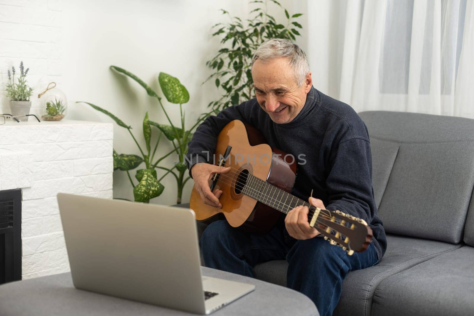 Portrait of senior man in headphones taking online guitar lesson looking at laptop screen. Retired male learning to play guitar watching webinar on computer at home by Andelov13