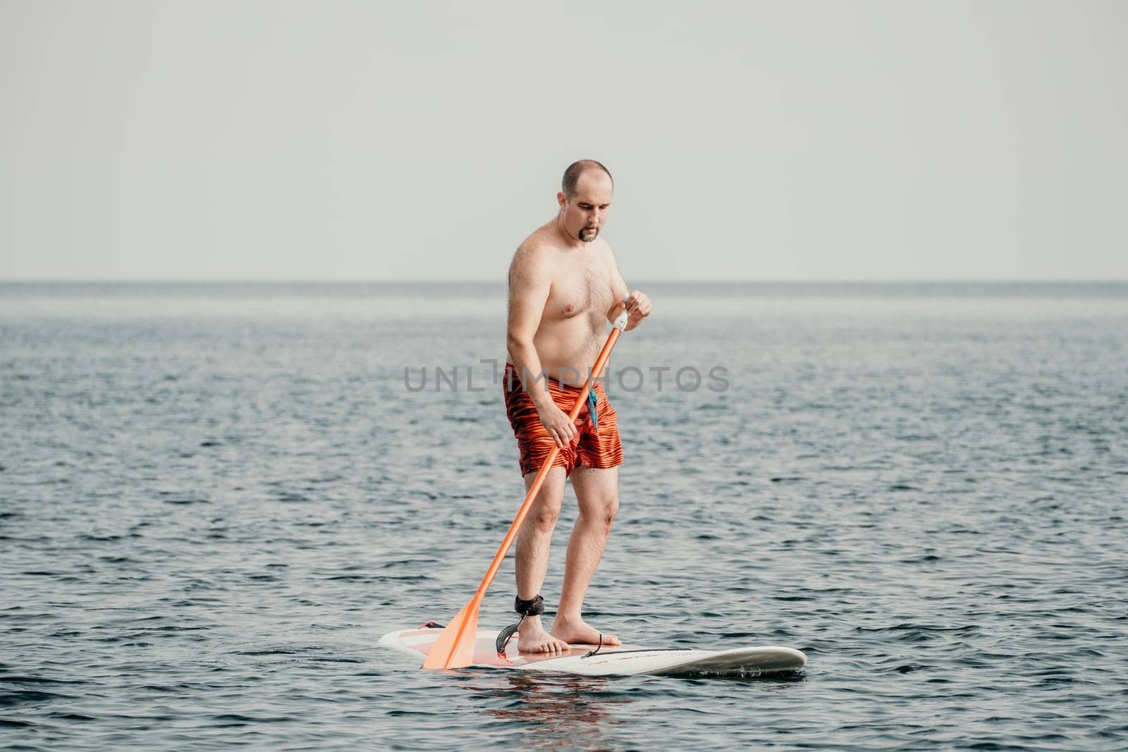 Active mature male paddler with his paddleboard and paddle on a sea at summer. Happy senior man stands with a SUP board. Stand up paddle boarding - outdor active recreation in nature