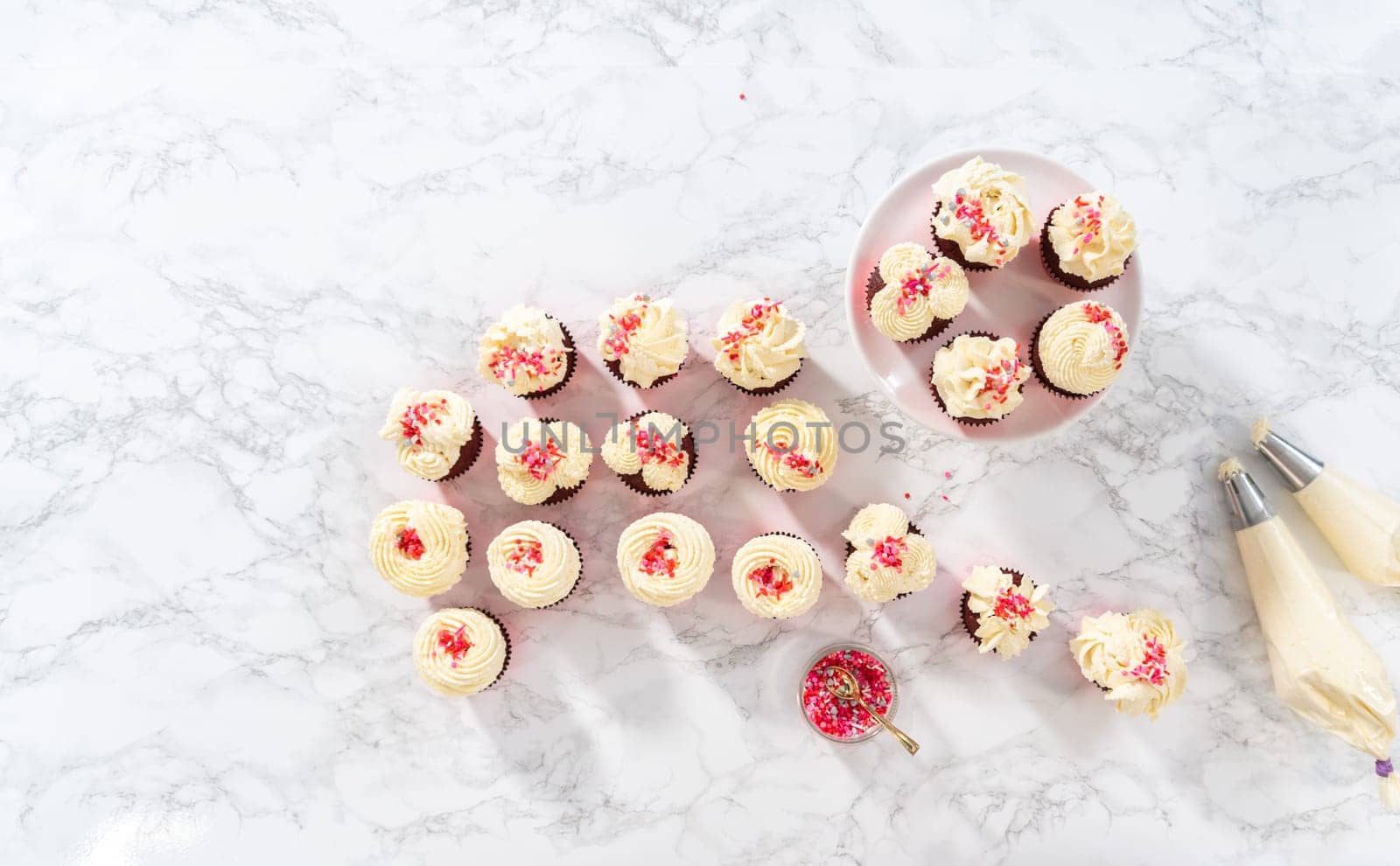 Flat lay. Piping white chocolate ganache frosting on top of red velvet cupcakes and topping with sprinkles.