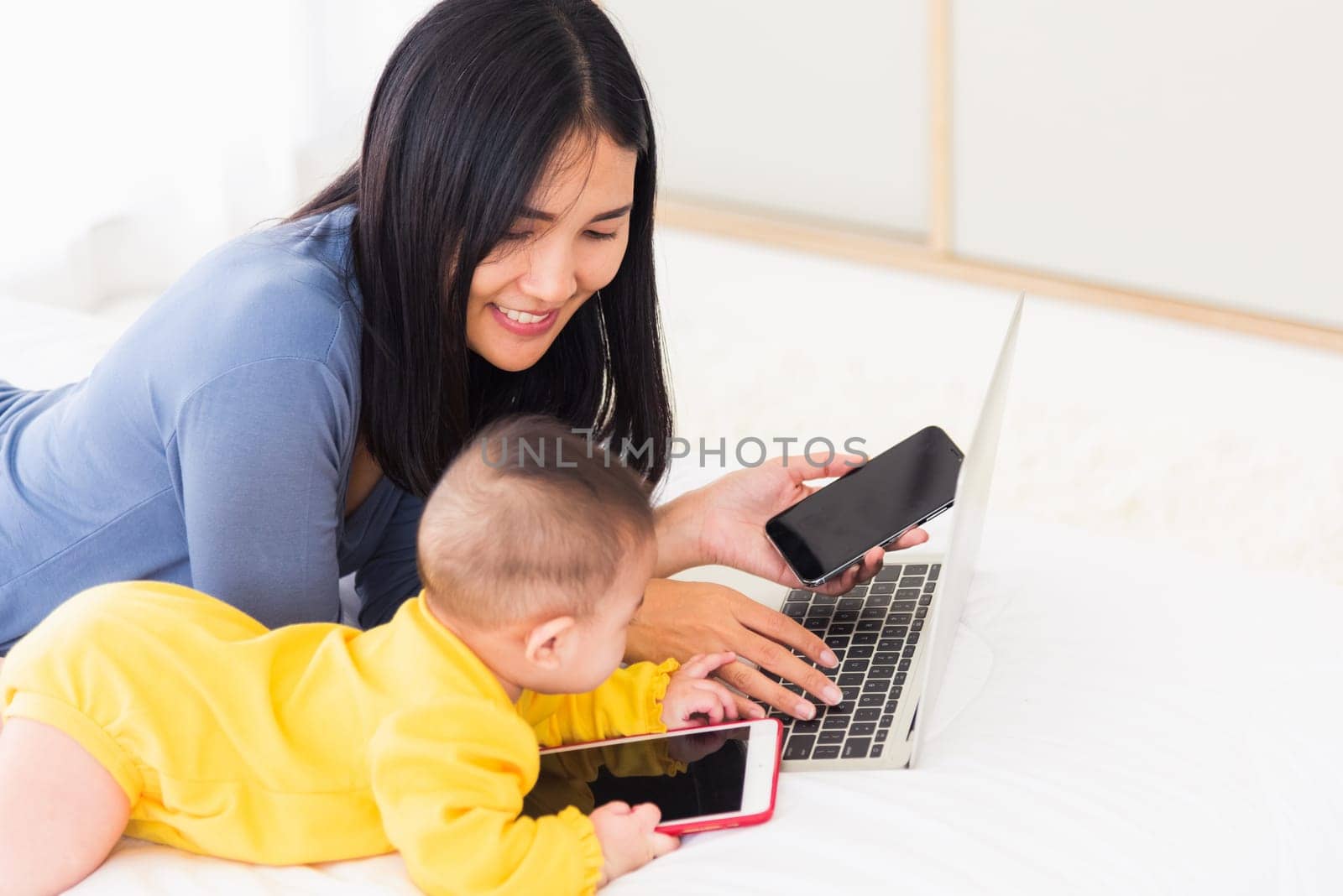 mother work from home busy on laptop computer and his baby daughter playing smartphone by Sorapop