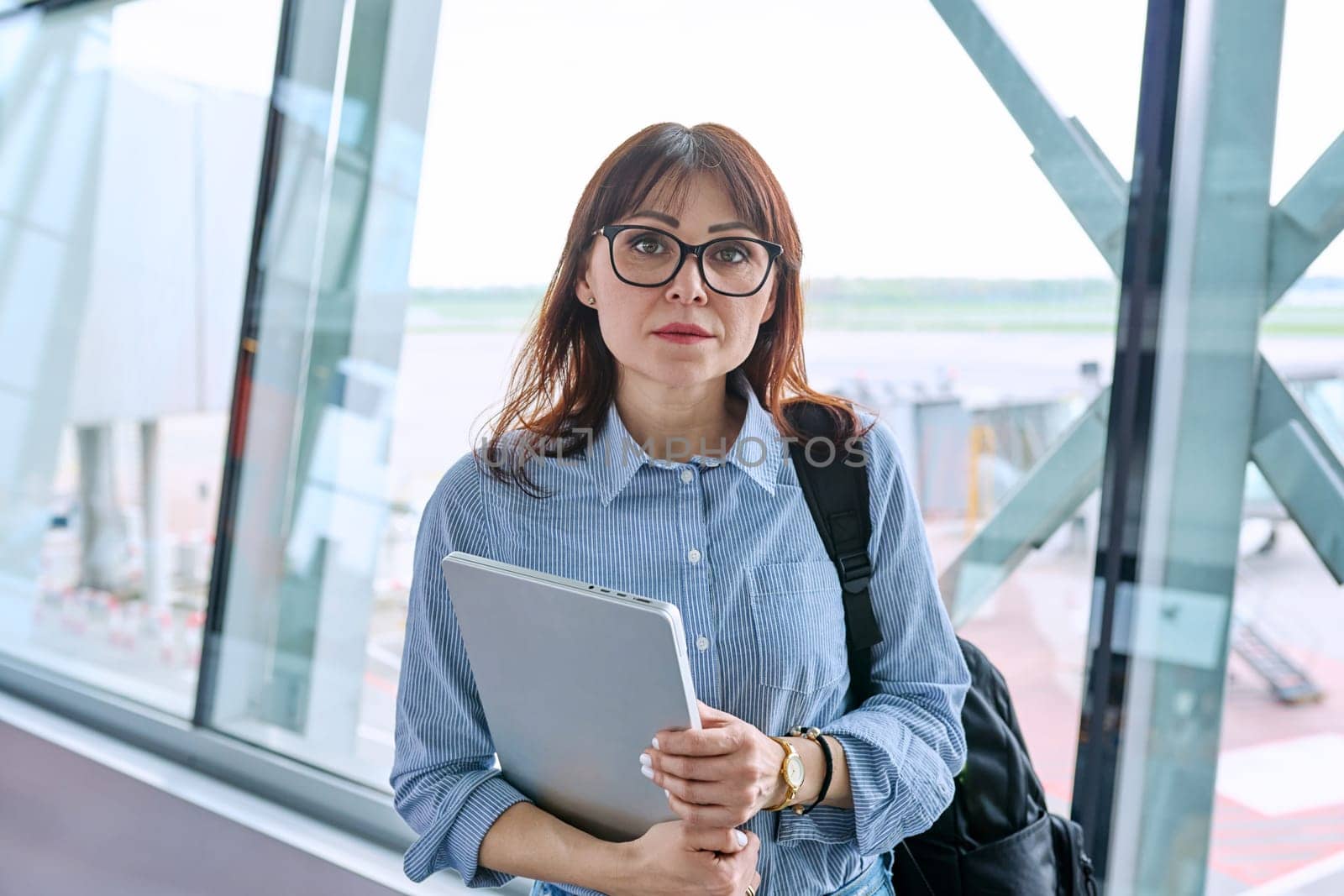 Middle aged woman with backpack waiting for airplane flight in airport terminal, holding laptop, looking at camera. Travel, business trip, passenger air transport concept