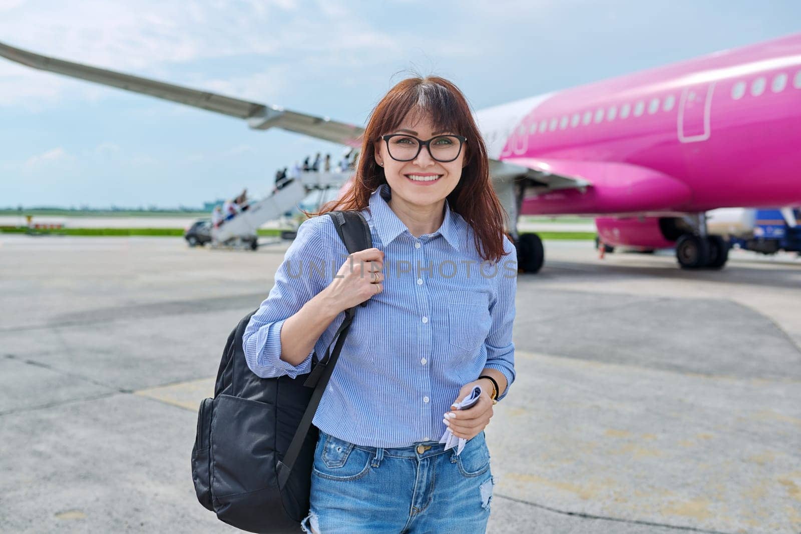 Outdoor portrait of smiling middle aged woman with hand luggage backpack looking at camera, passengers boarding plane background. Travel, business trip, air transport concept