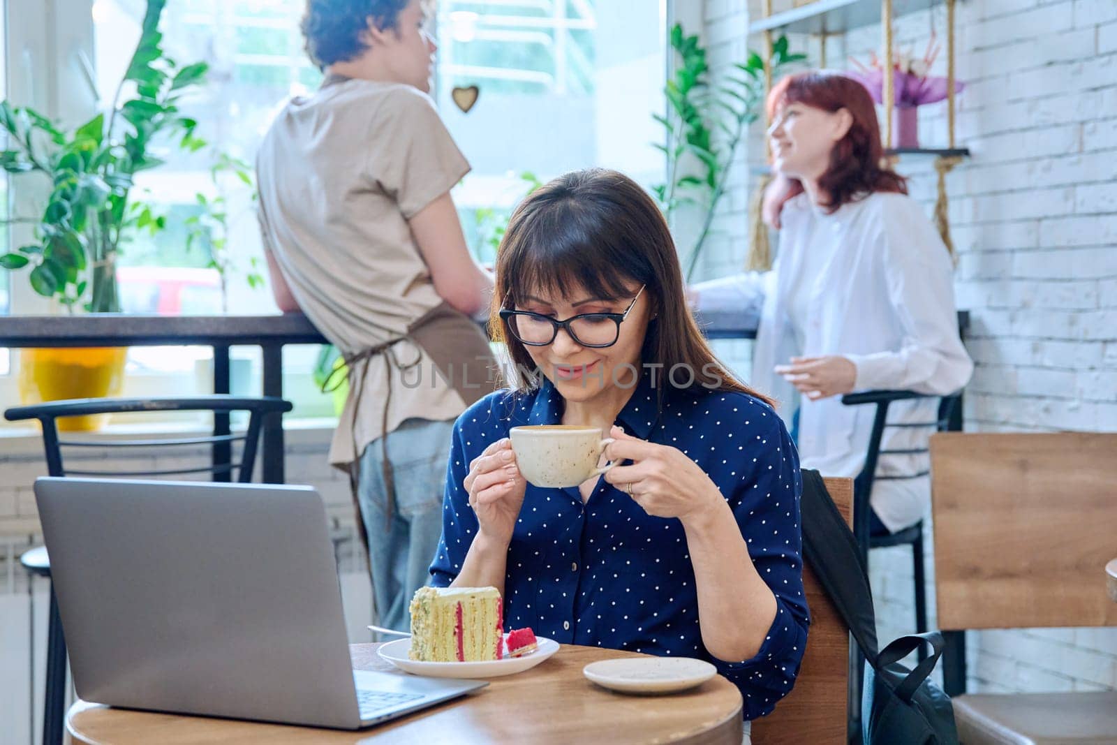 Middle aged woman in bakery cafeteria with cup of coffee and dessert cake, sitting at table, with laptop. Lunch break, food, lifestyle, mature 40s people concept