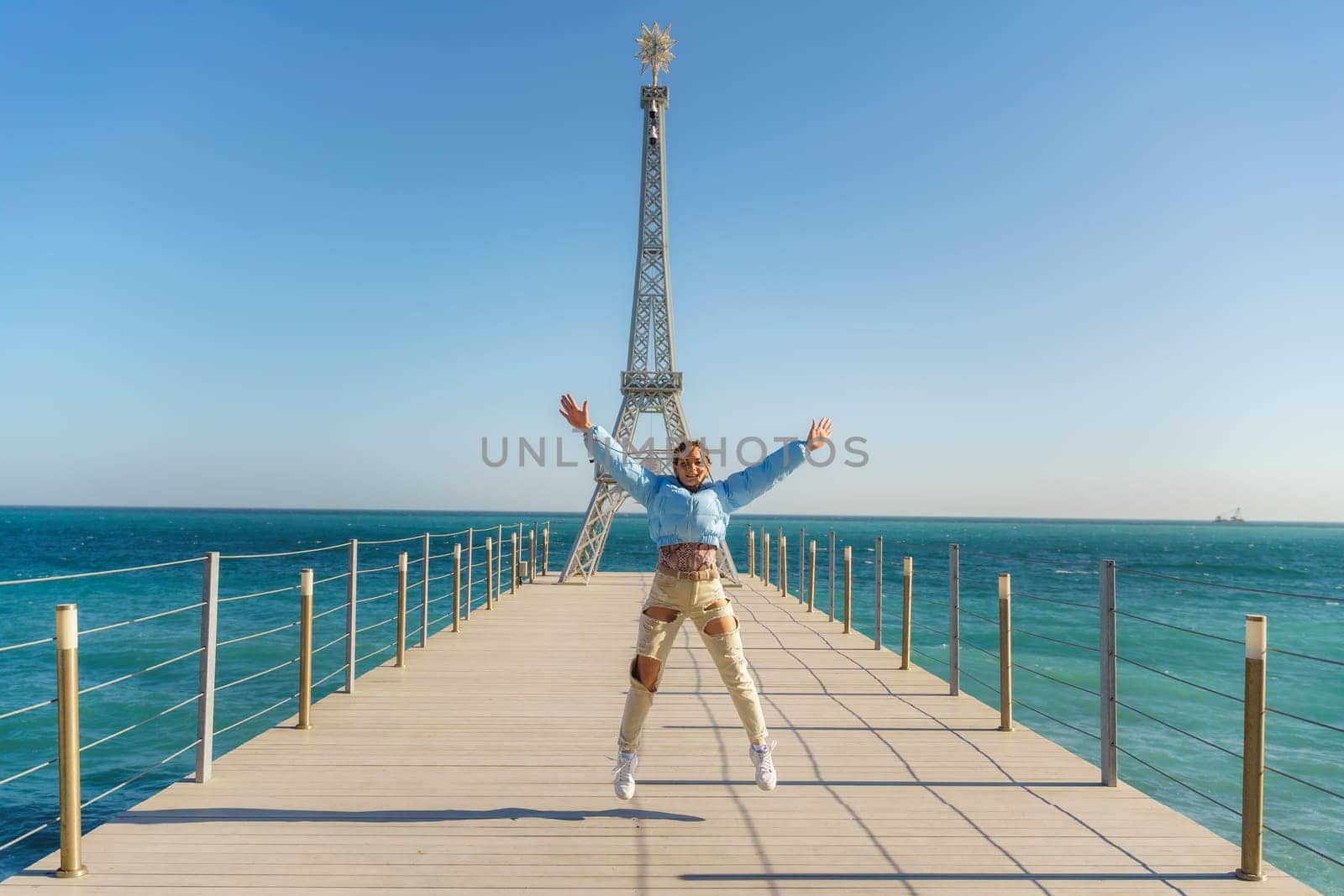 Large model of the Eiffel Tower on the beach. A woman walks along the pier towards the tower, wearing a blue jacket and white jeans