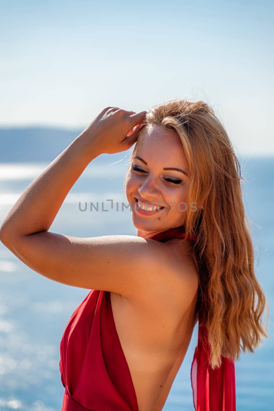 Smiling young woman in a red dress looks at the camera. A beautiful tanned girl enjoys her summer holidays at the sea. Portrait of a stylish carefree woman laughing at the ocean