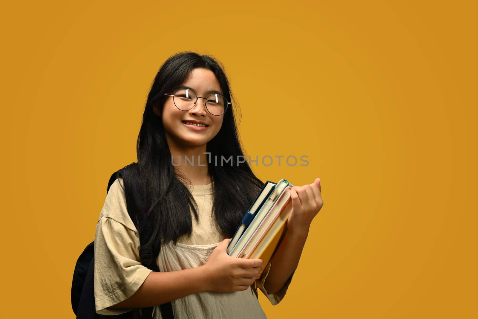 Smiling teenager schoolgirl with backpack holding books standing on yellow background. Back to school concept..