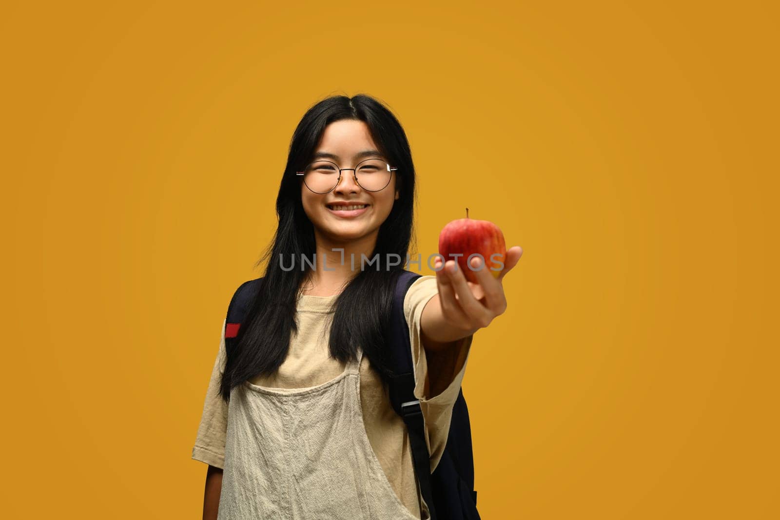 Happy schoolgirl wearing glasses holding an apple on yellow background. Educational, learning and back to school concept.