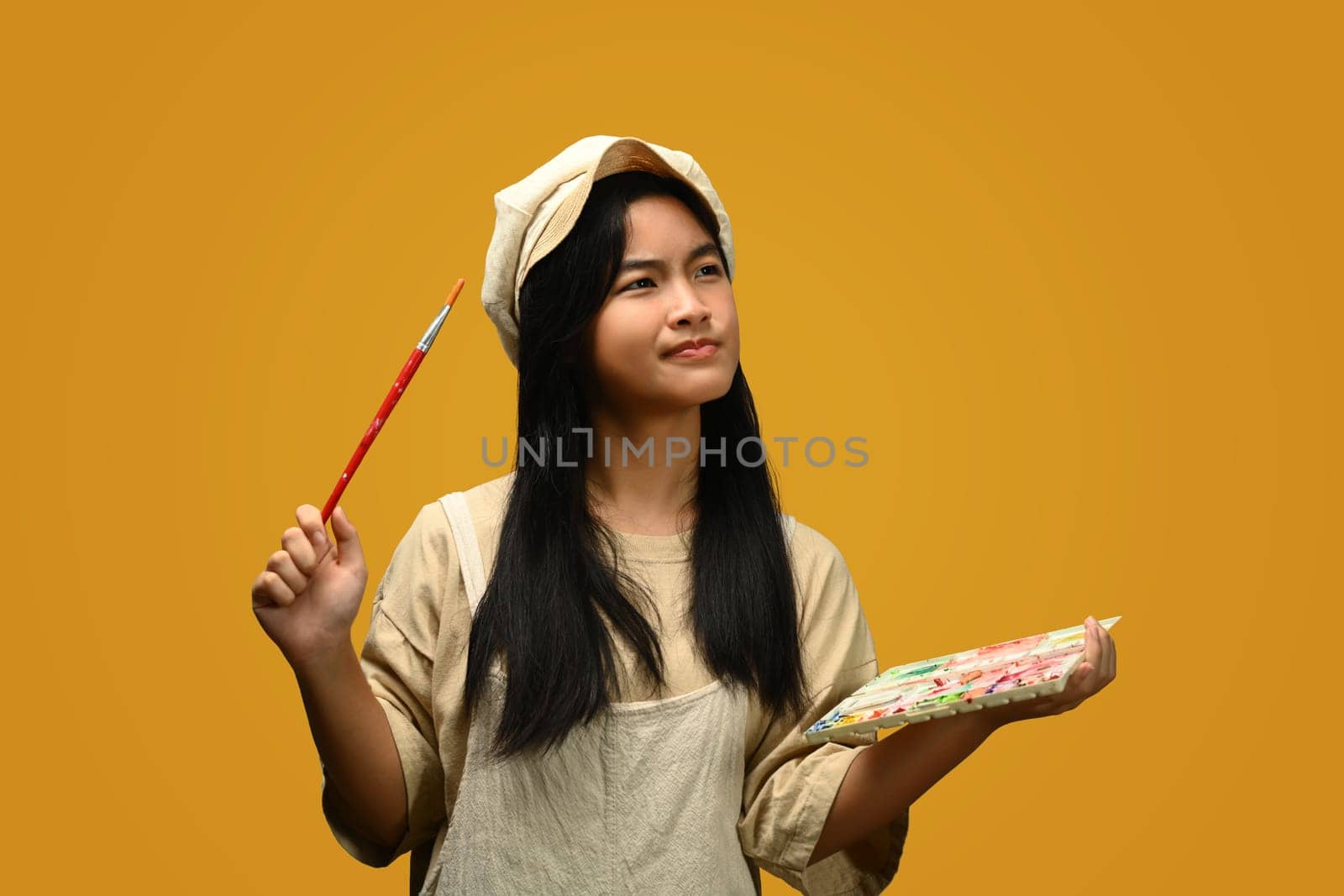 Cheerful teenage Asian girl holding palette and paint brushes on yellow background. Art, creativity and education concept.