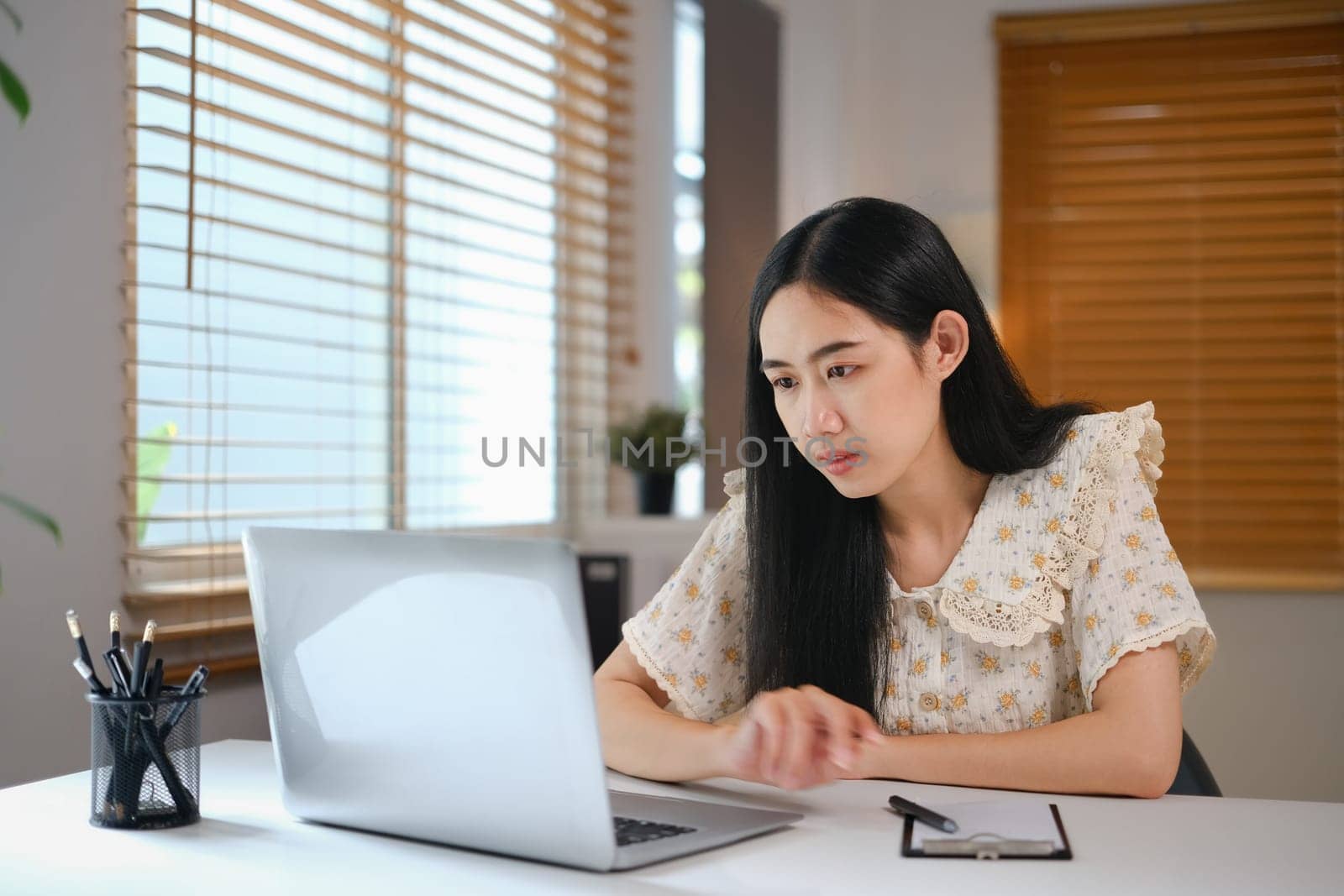 Focused young Asian woman looking at laptop screen, working or studying online at home.