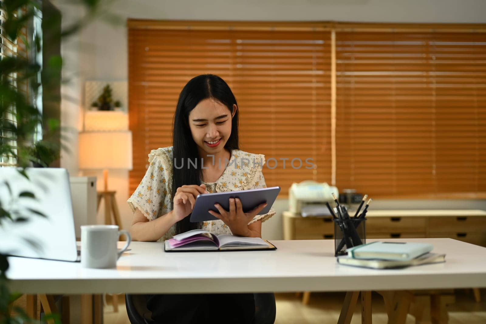 Pleasant young woman in casual clothes using digital tablet, working in cozy home office..