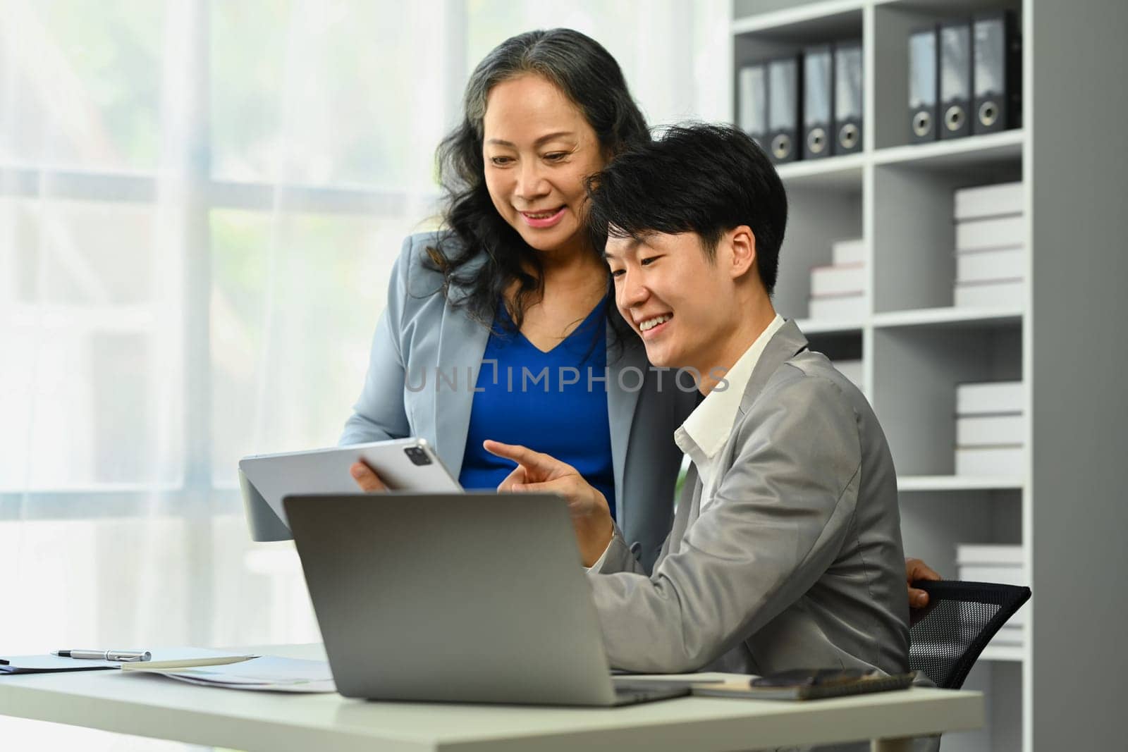 Positive mature businesswoman helping and explaining work detail to young businessman.