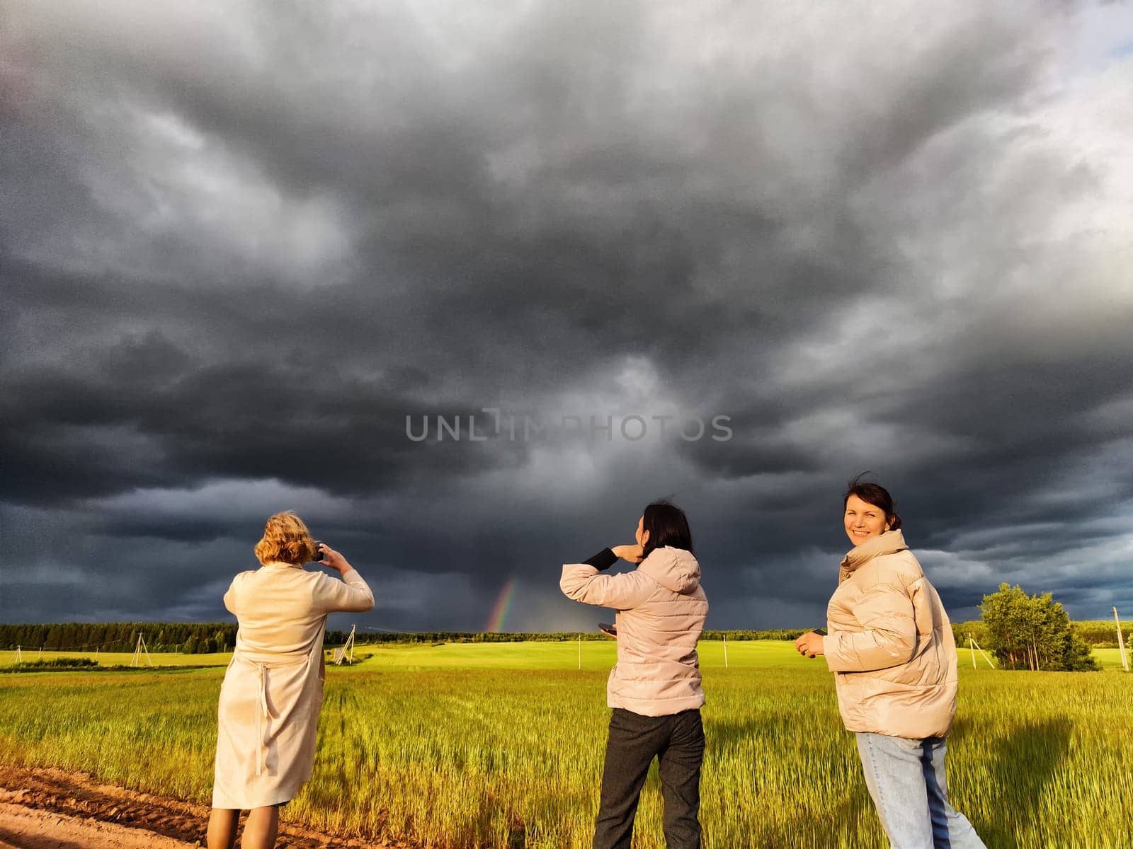 Tourist girls take pictures of a rainbow and dramatic, gloomy clouds over a field on a spring, summer, autumn day. The concept of the beauty of nature and travel by keleny