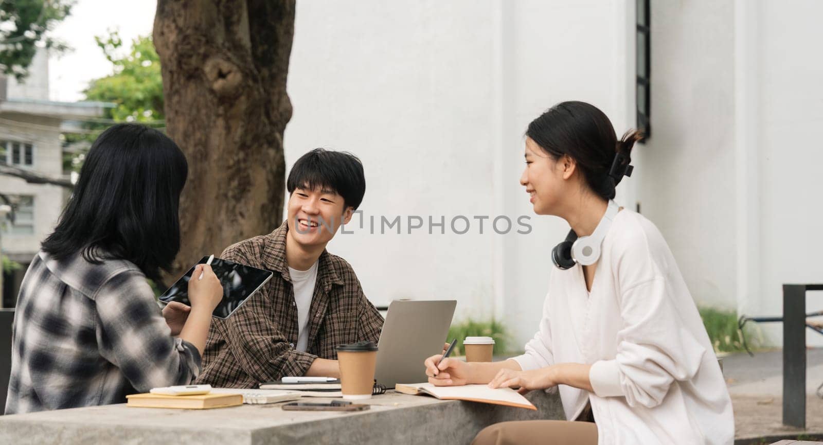 Group of happy young Asian college students sitting on a on the cement table, looking at a laptop screen, discussing and brainstorming on their university project together by nateemee