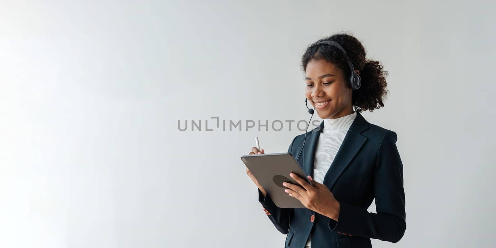 Call center operator in wireless headset talking with customer on tablet, African woman in headphones with microphone consulting client on phone in customer support service in business center.