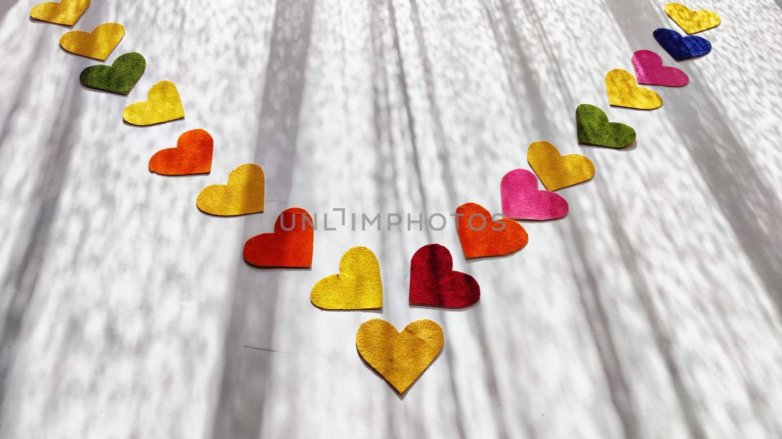 Multicolored hearts made of rough velvet paper on white surface with stripes of shadow and light from the sun. Abstract background, texture, copy space