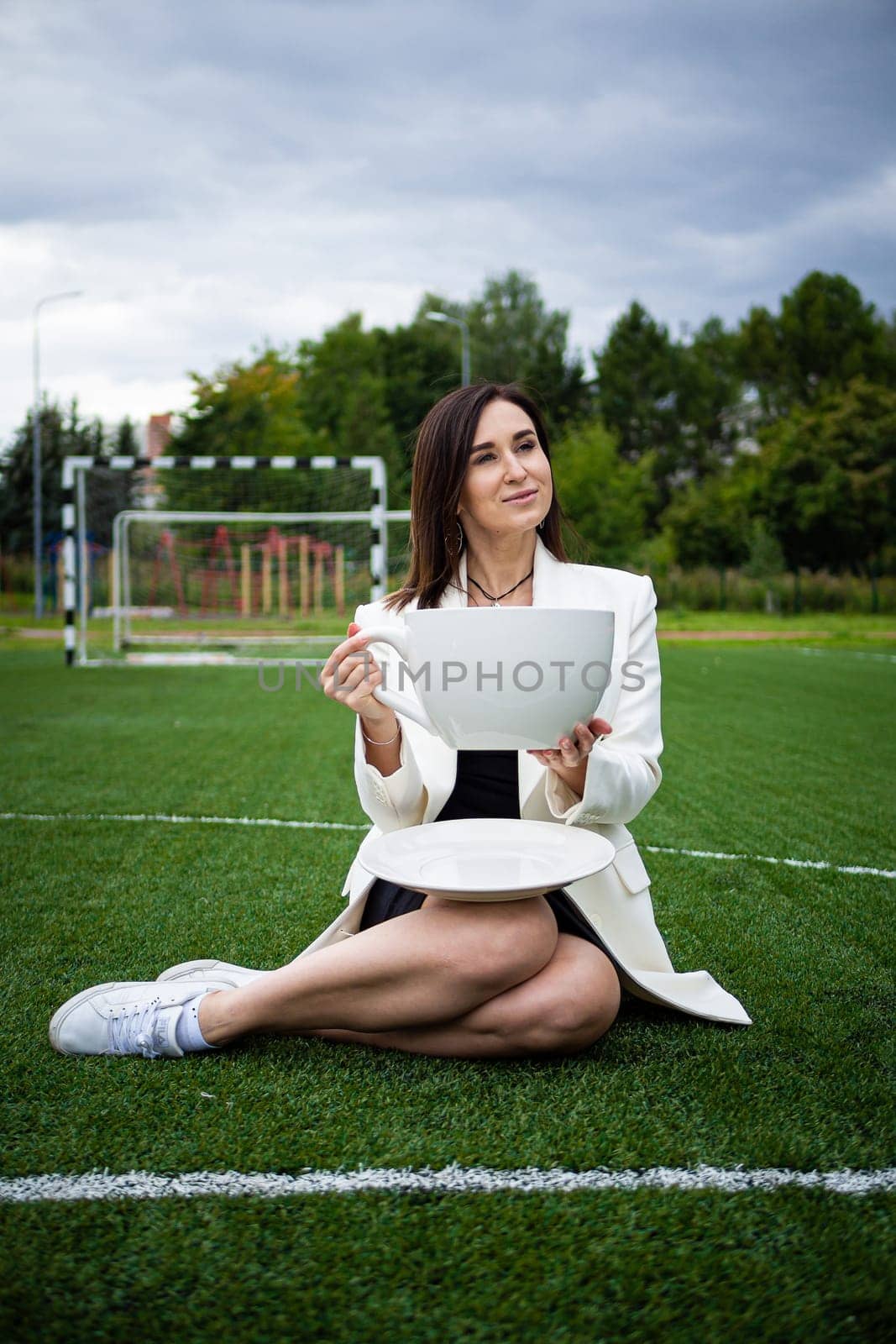 A business woman with a large cup, sitting on a green lawn in the park. The concept of an office worker on a picnic.