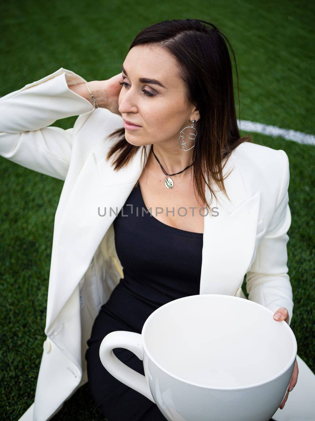 A business woman with a large cup, sitting on a green lawn in the park. The concept of an office worker on a picnic.