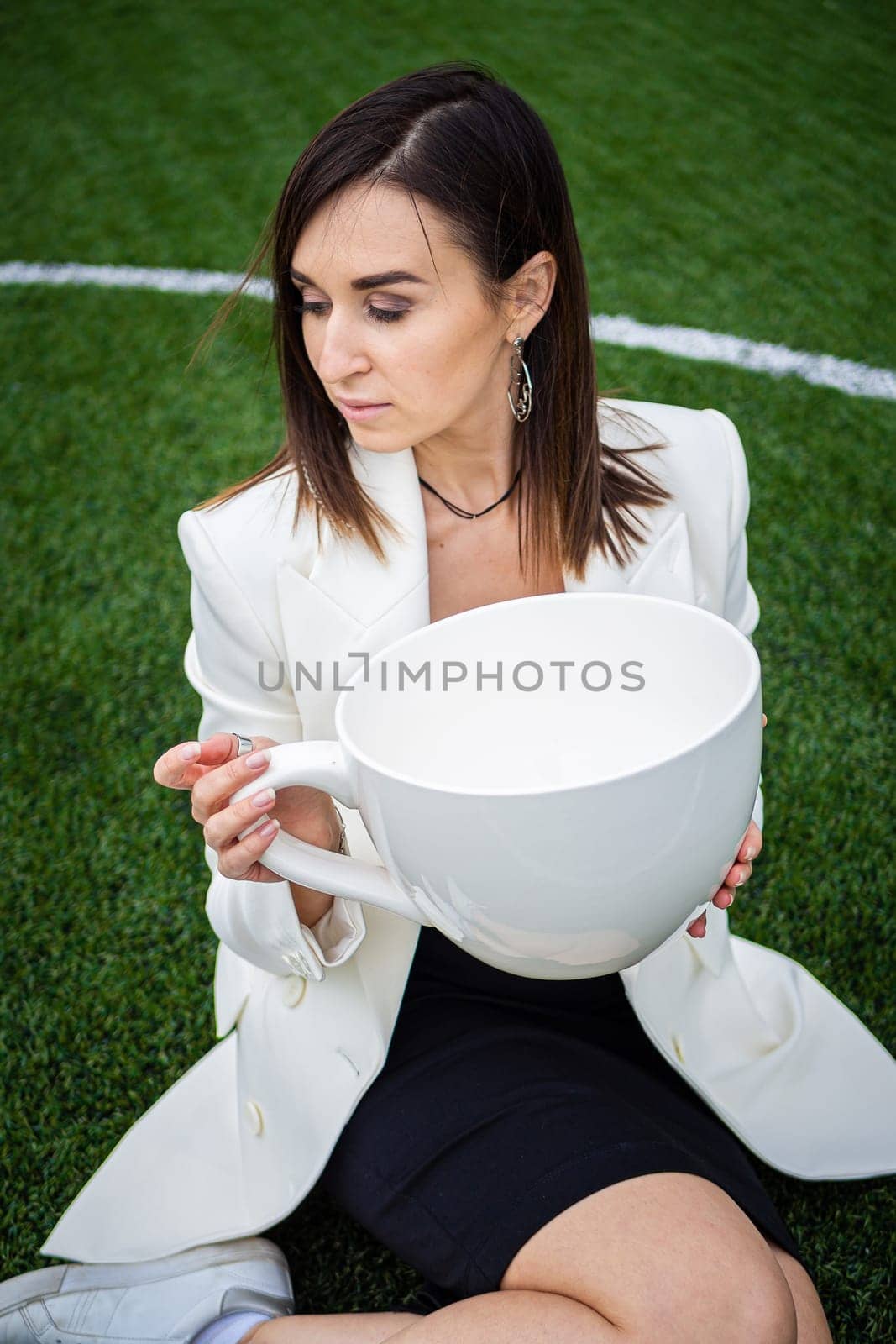 A business woman with a large cup, sitting on a green lawn in the park. The concept of an office worker on a picnic.