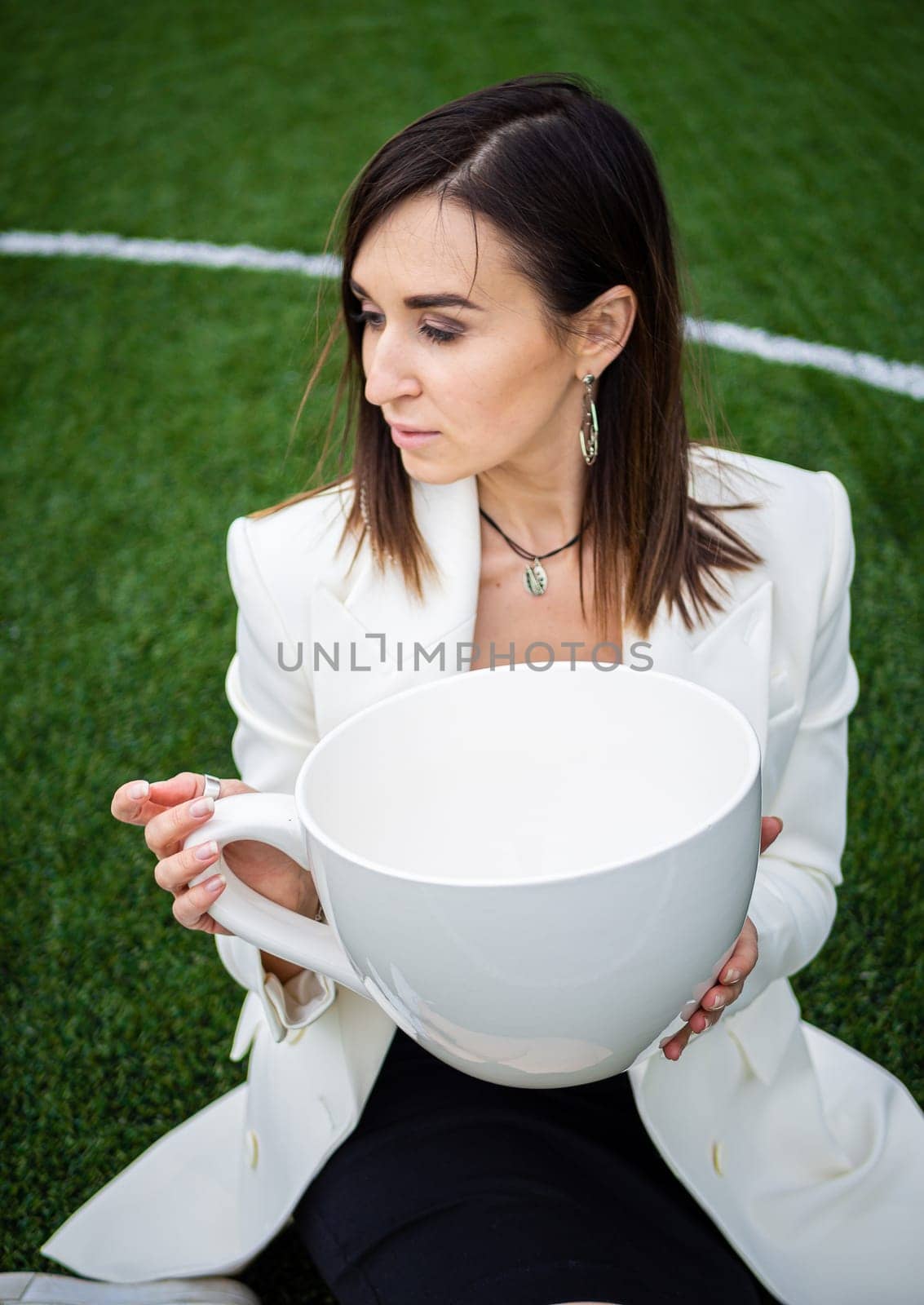 A business woman with a large cup, sitting on a green lawn in the park. by Evgenii_Leontev