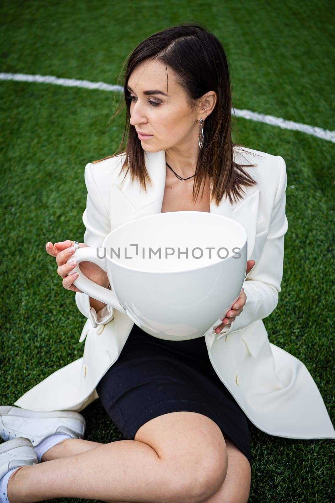 A business woman with a large cup, sitting on a green lawn in the park. The concept of an office worker on a picnic.