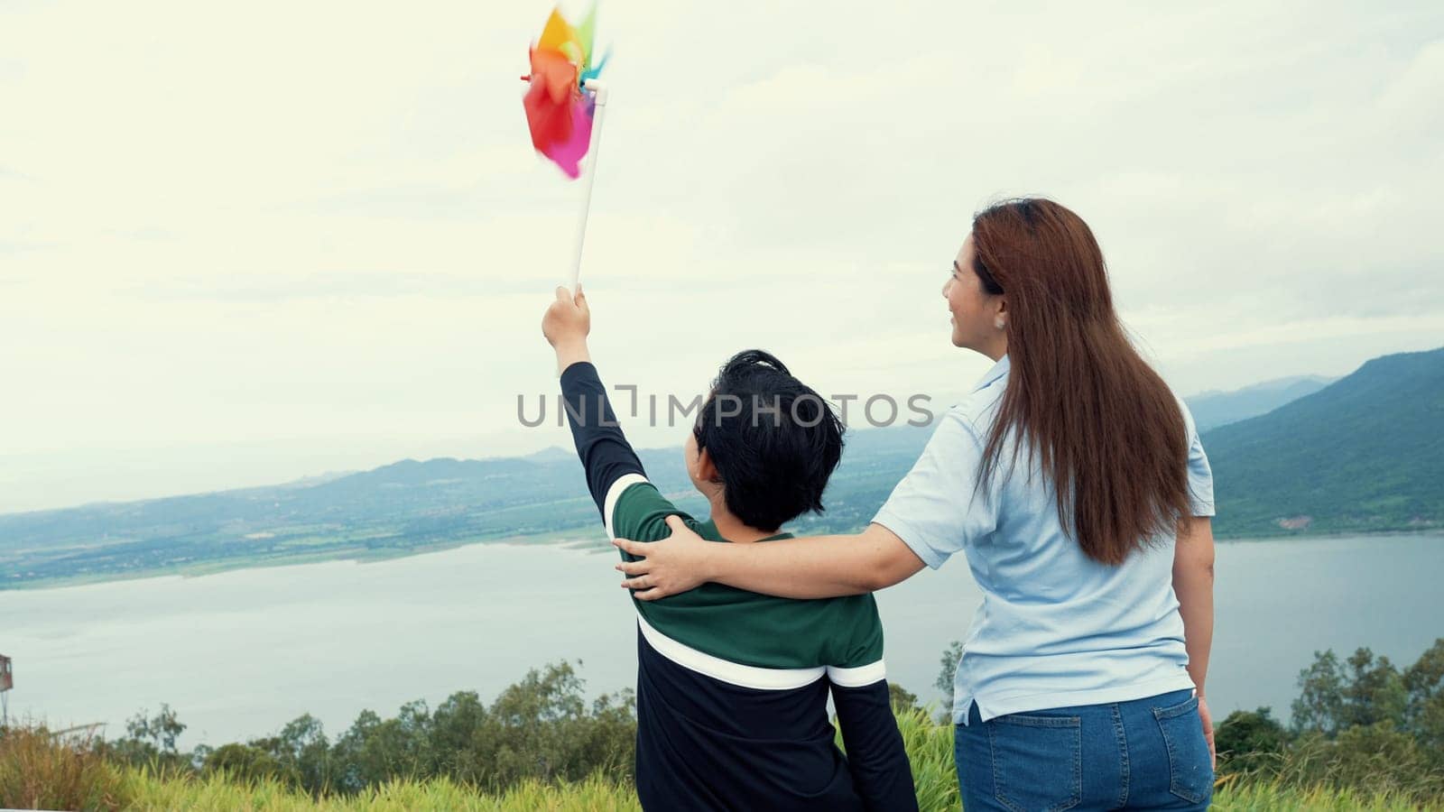 A progressive woman and her son are on vacation, enjoying the natural beauty of a lake at the bottom of a hill while the boy carries a toy windmill.