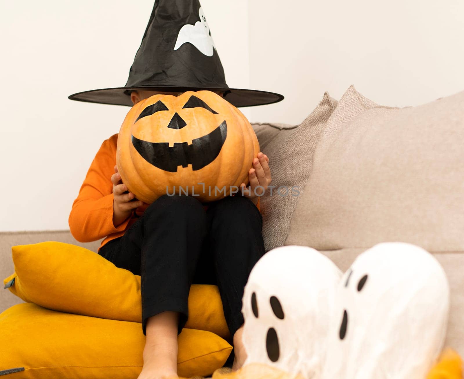 Halloween. Concept. A boy in an orange sweater and a haunted wizard's hat, with a pumpkin instead of a face, sits cheerfully in a home interior. Close-up