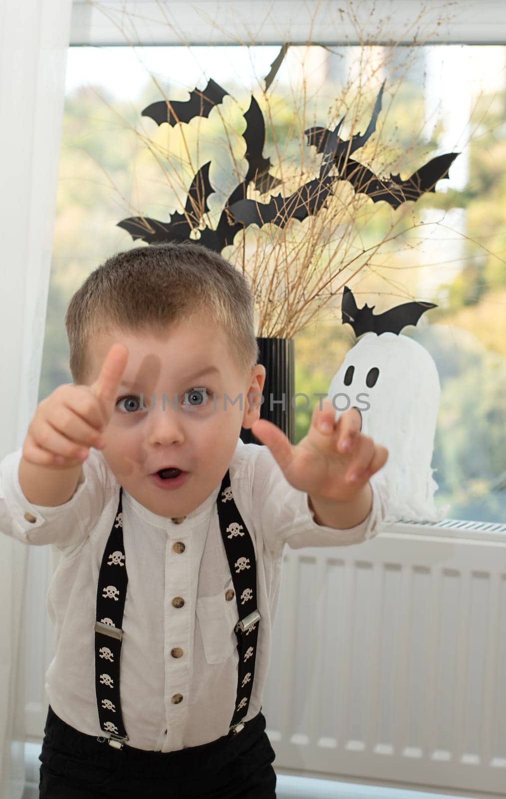 Halloween concept. A little boy, with emotional facial expressions, in a white shirt and black suspenders with images of skulls against the background of a window with a vase of dry branches and black paper bats. Close-up