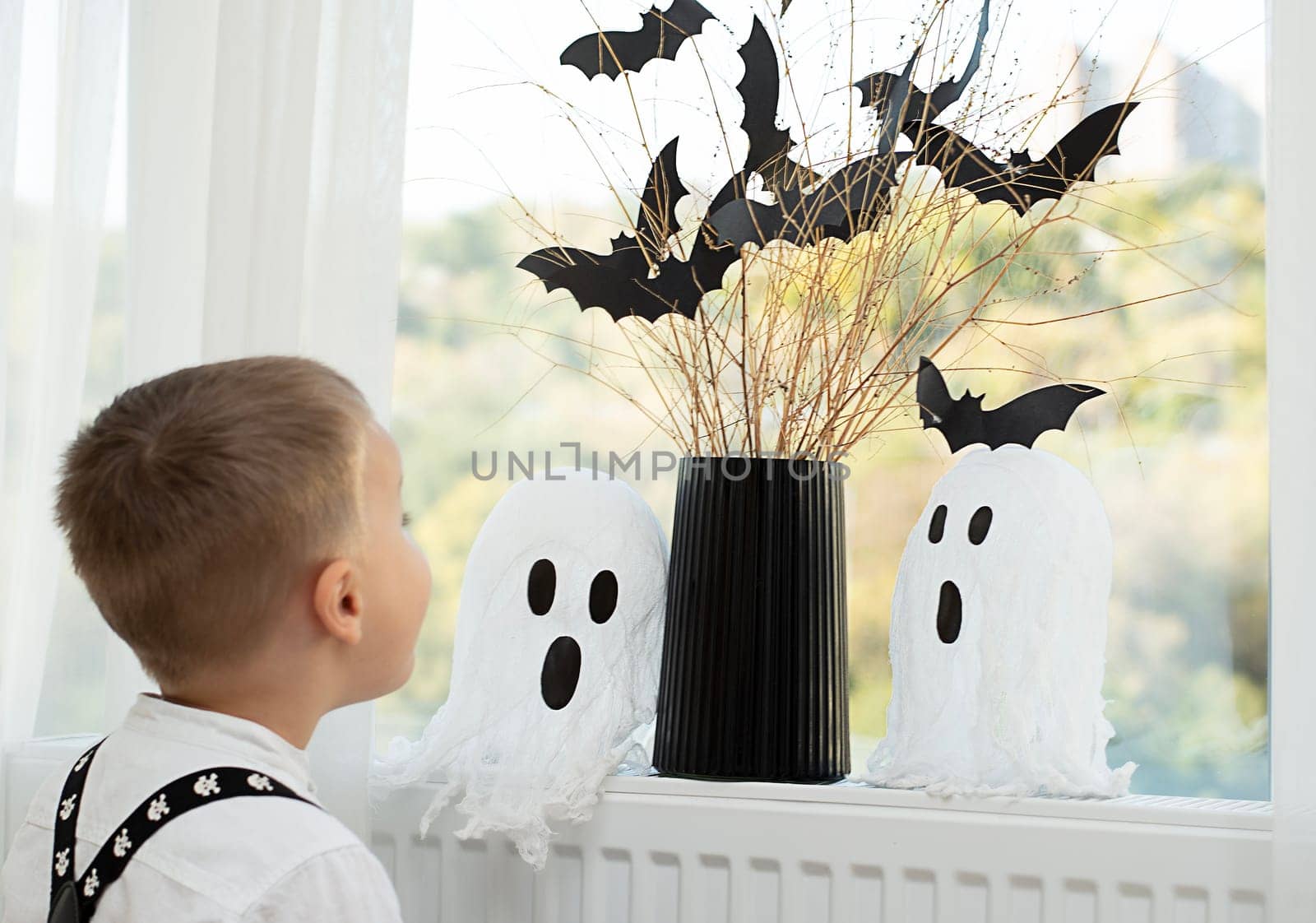 Halloween concept. A little boy, with emotional facial expressions, in a white shirt and black suspenders with images of skulls against the background of a window with a vase of dry branches and black paper bats. Close-up