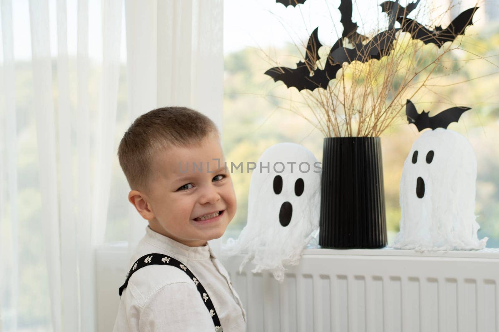 Halloween concept. A little boy, with emotional facial expressions, in a white shirt and black suspenders with images of skulls against the background of a window with a vase of dry branches and black paper bats. Close-up