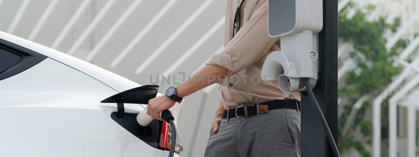 Young man recharge electric car's battery from charging station in city commercial parking lot. Rechargeable EV car for sustainable environmental friendly urban travel. Panorama Expedient