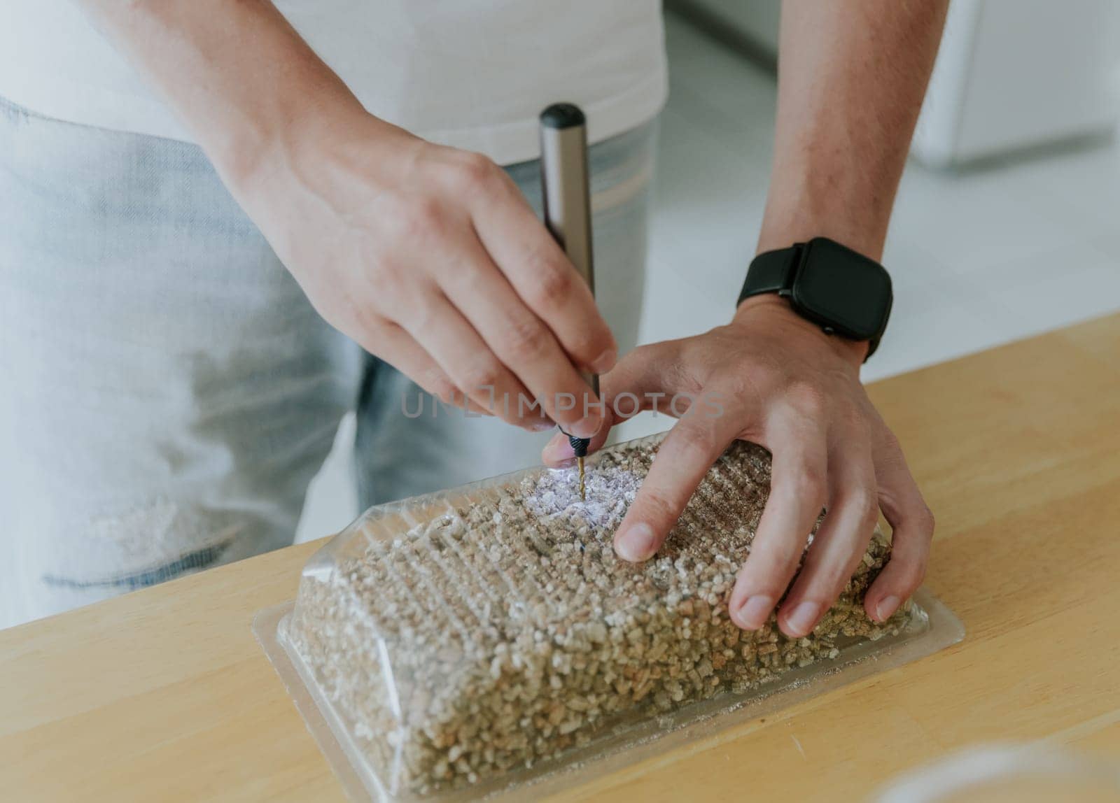 One young Caucasian man drills a plastic transparent box with filler for planting wheat with a minidrill, standing in the kitchen at the table, close-up side view.